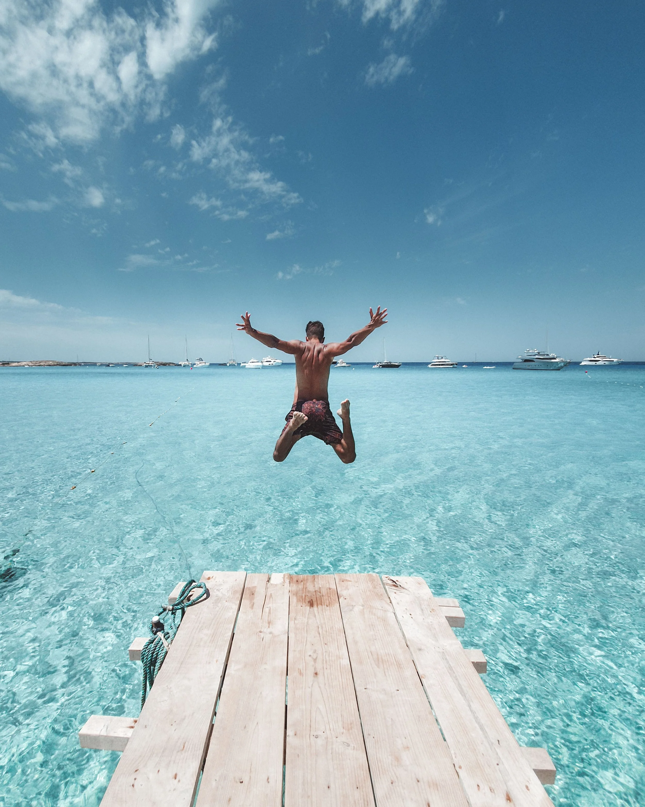 Person jumping off a wooden dock into clear blue water, with boats and yachts anchored in the background and a sky with scattered clouds.