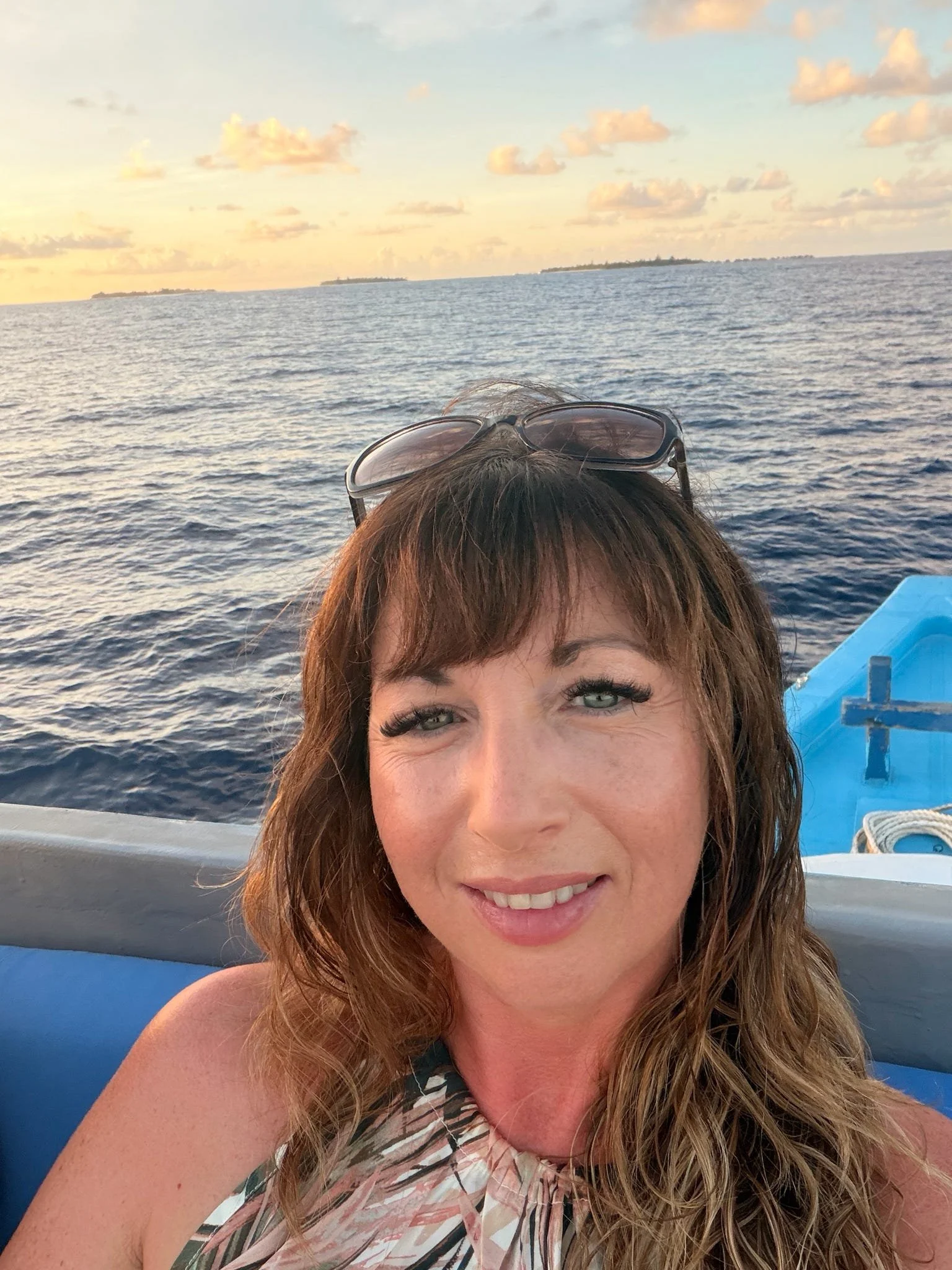 A woman with wavy brown hair and sunglasses on her head, smiling, on a boat with water and distant land in the background during sunset.