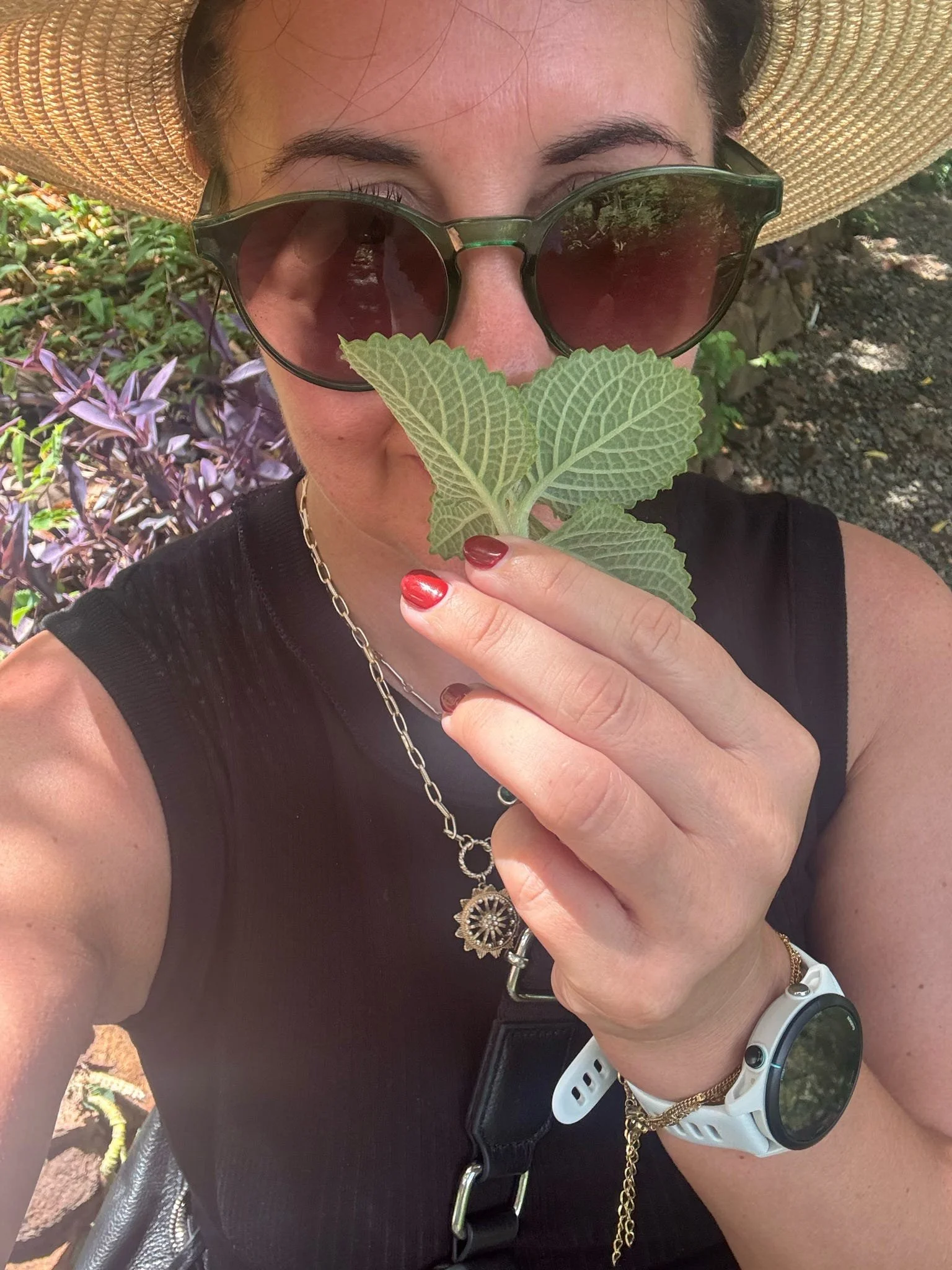 Close-up of a woman wearing a large straw hat, dark sunglasses, a black sleeveless top, and gold jewelry, holding a green leaf in front of her face outdoors.