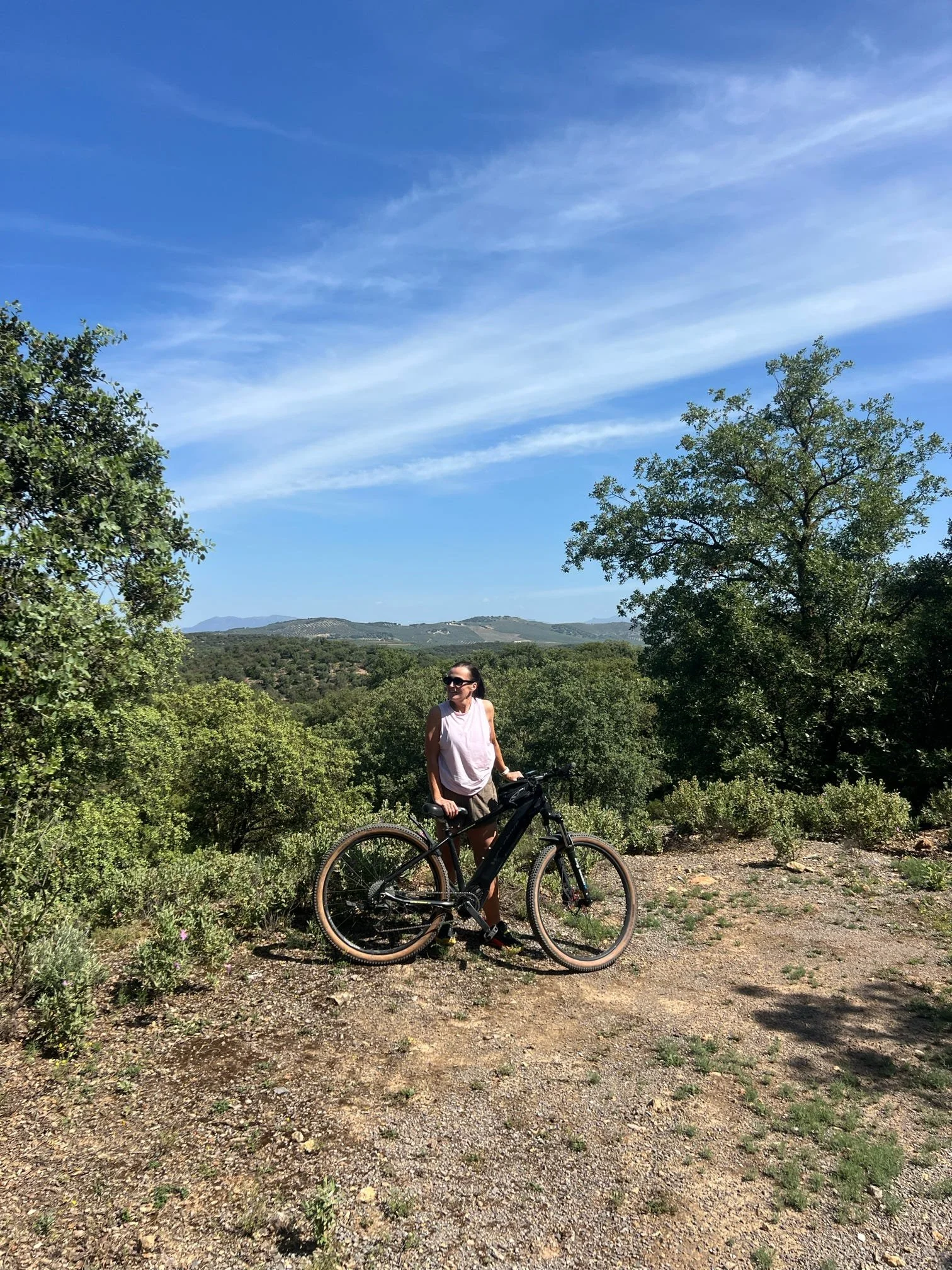 A woman standing with a bicycle on a dirt trail in a hilly, green landscape under a bright blue sky with wispy clouds.