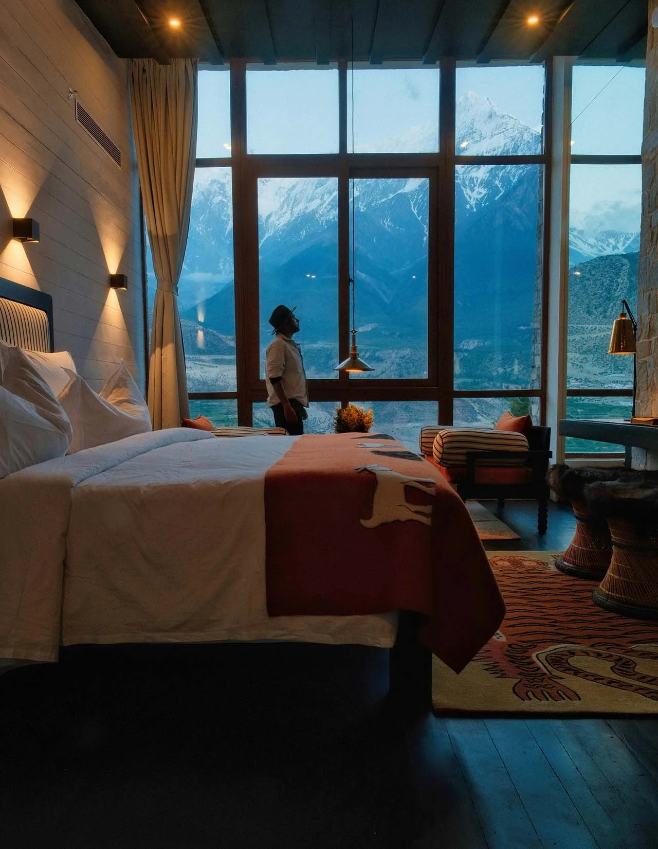 A hotel room with a large window showing snow-capped mountains. Inside, there is a bed with white and orange bedding, a person standing by the window, and sitting chairs with striped cushions.