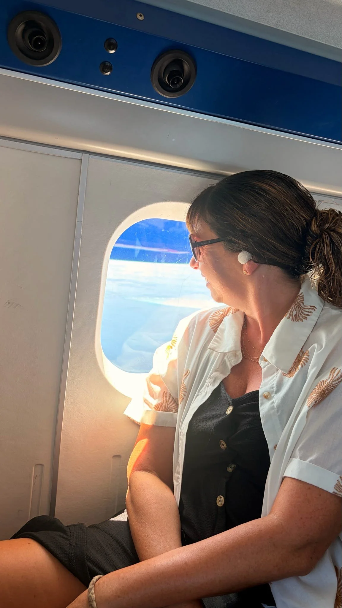 A woman with glasses sits by an airplane window, looking outside at the sky.