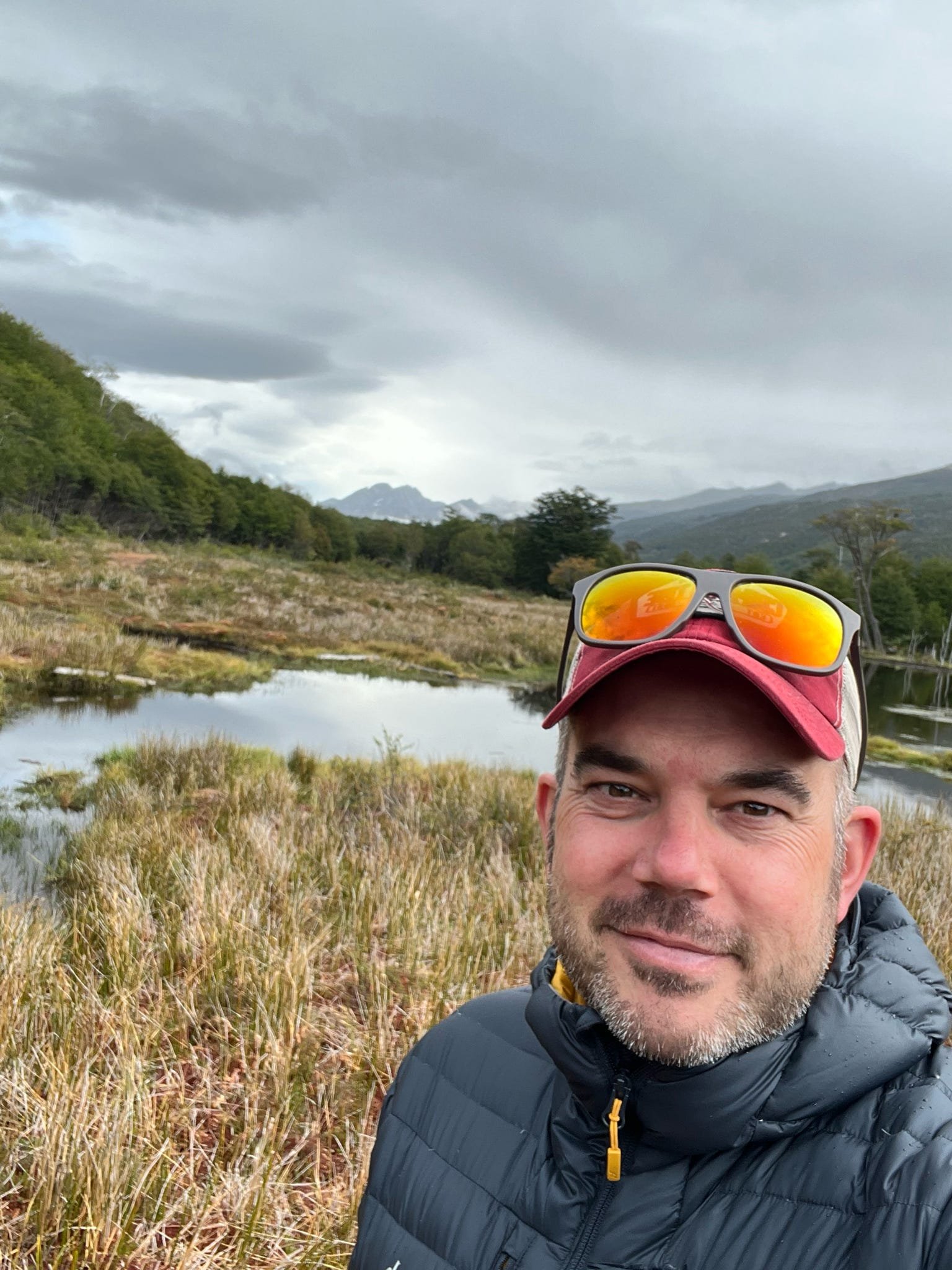 A man with short dark hair, beard, and wearing a black puffy jacket and a red cap with reflective sunglasses on top, takes a selfie in a natural landscape with tall grasses, a river, and distant mountains under cloudy sky.
