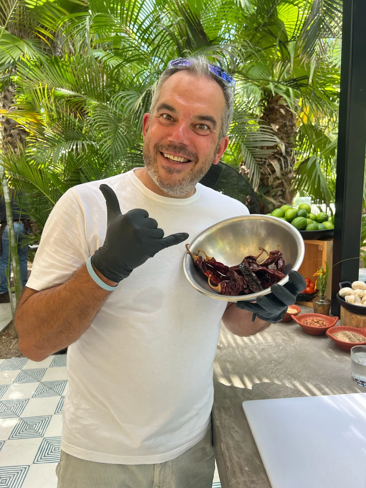 A smiling man with gray hair and a beard, wearing a white t-shirt, black gloves, and sunglasses on his head, holding a metal bowl with dried red chili peppers and making a shaka sign with his right hand. He is standing in front of lush green tropical