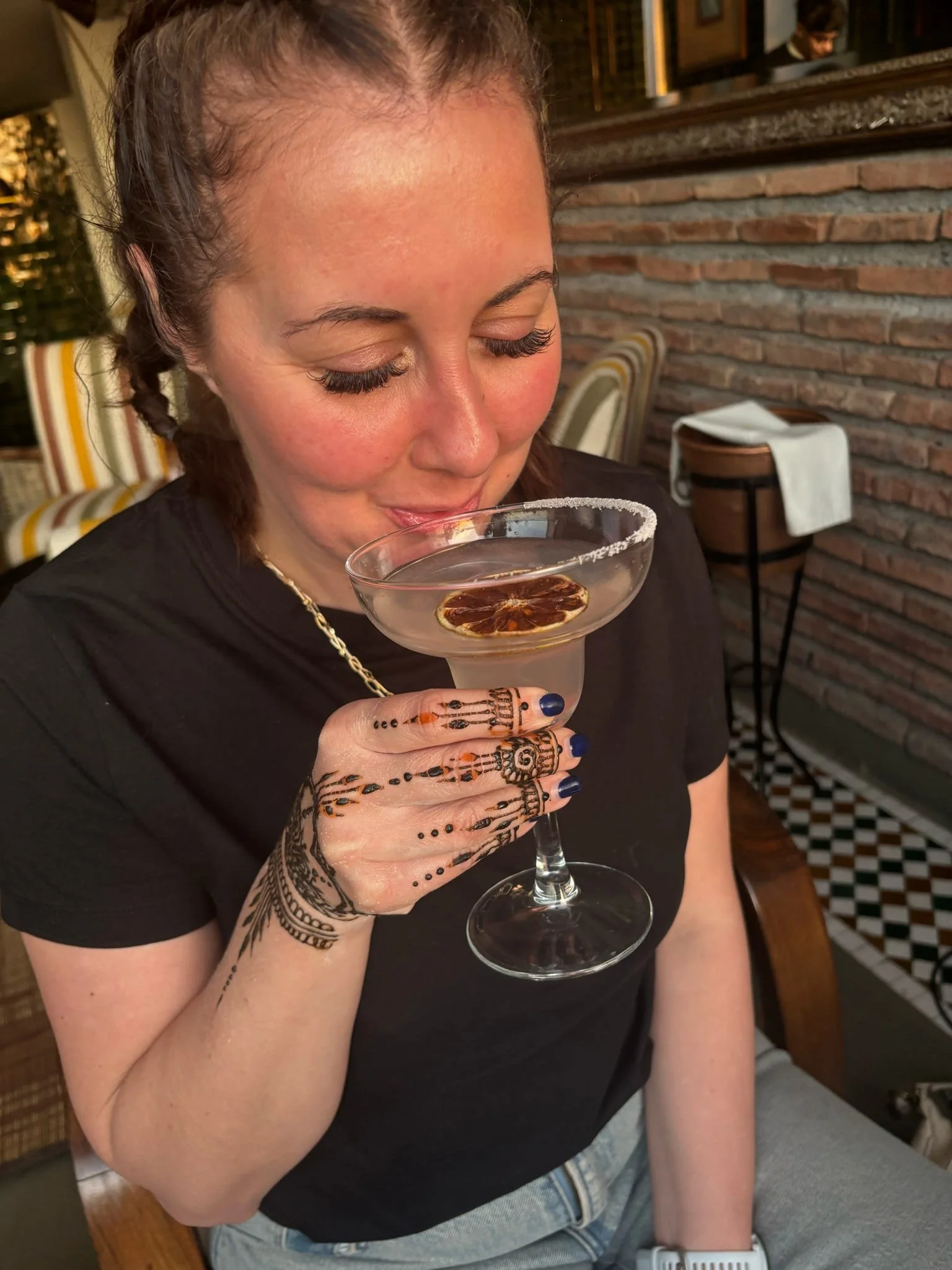 A woman with henna tattoos on her hand holding a cocktail glass with a dark-colored beverage garnished with a dried lemon slice, smiling and looking down with closed eyes.