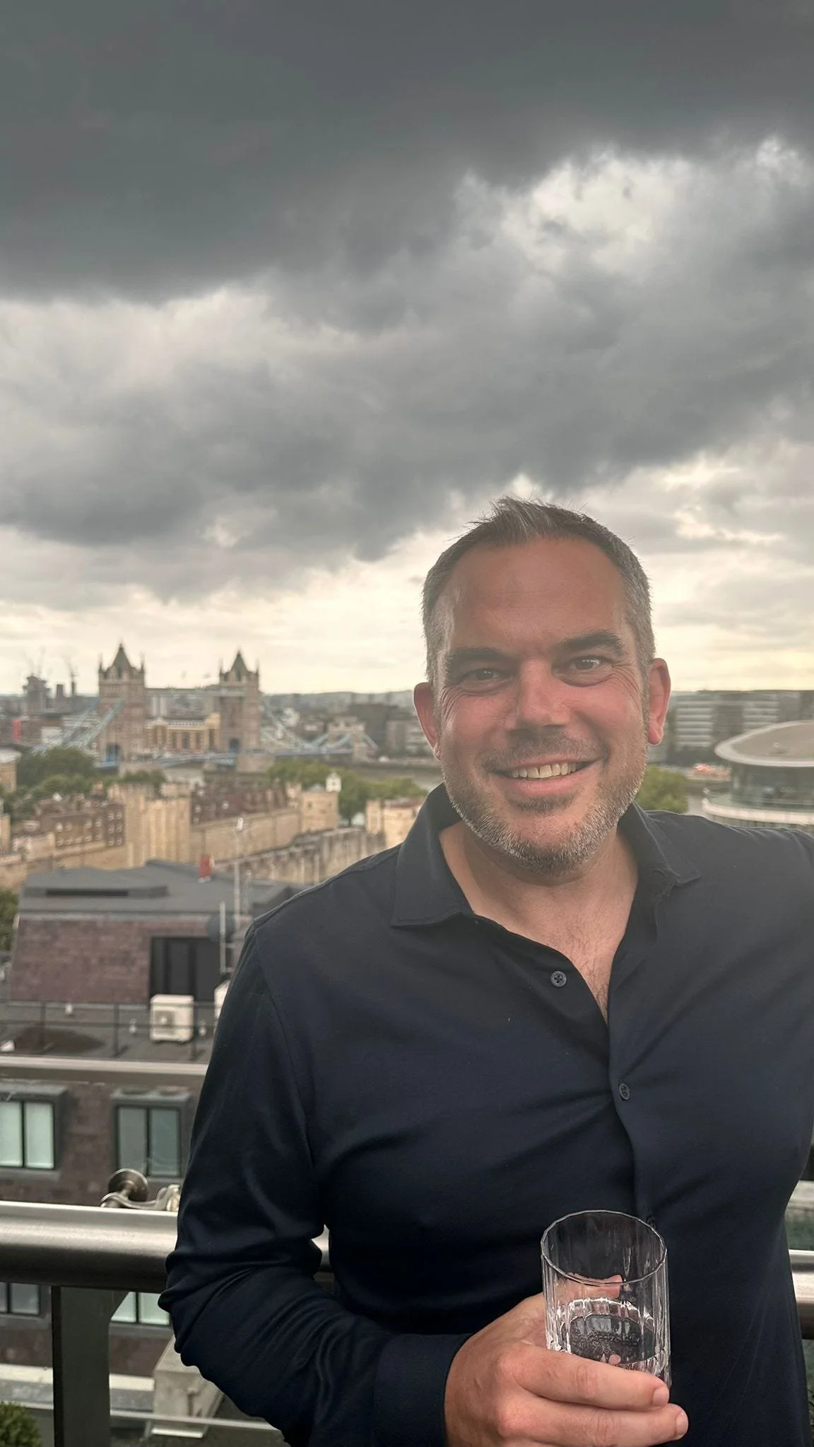 A smiling man with a beard and short hair, wearing a black shirt, is holding a glass and standing outdoors on a balcony with a city skyline and cloudy sky in the background.