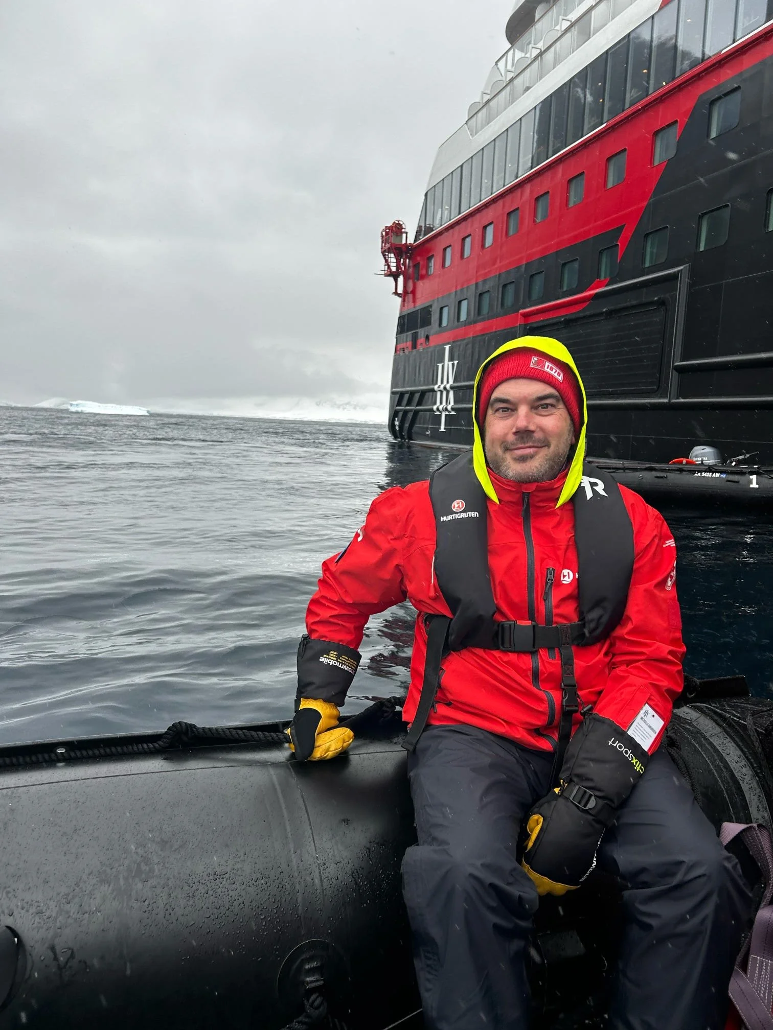 A man in a red jacket and a red beanie hat sitting on a boat during a cold, overcast day, with a large cruise ship in the background on icy waters.