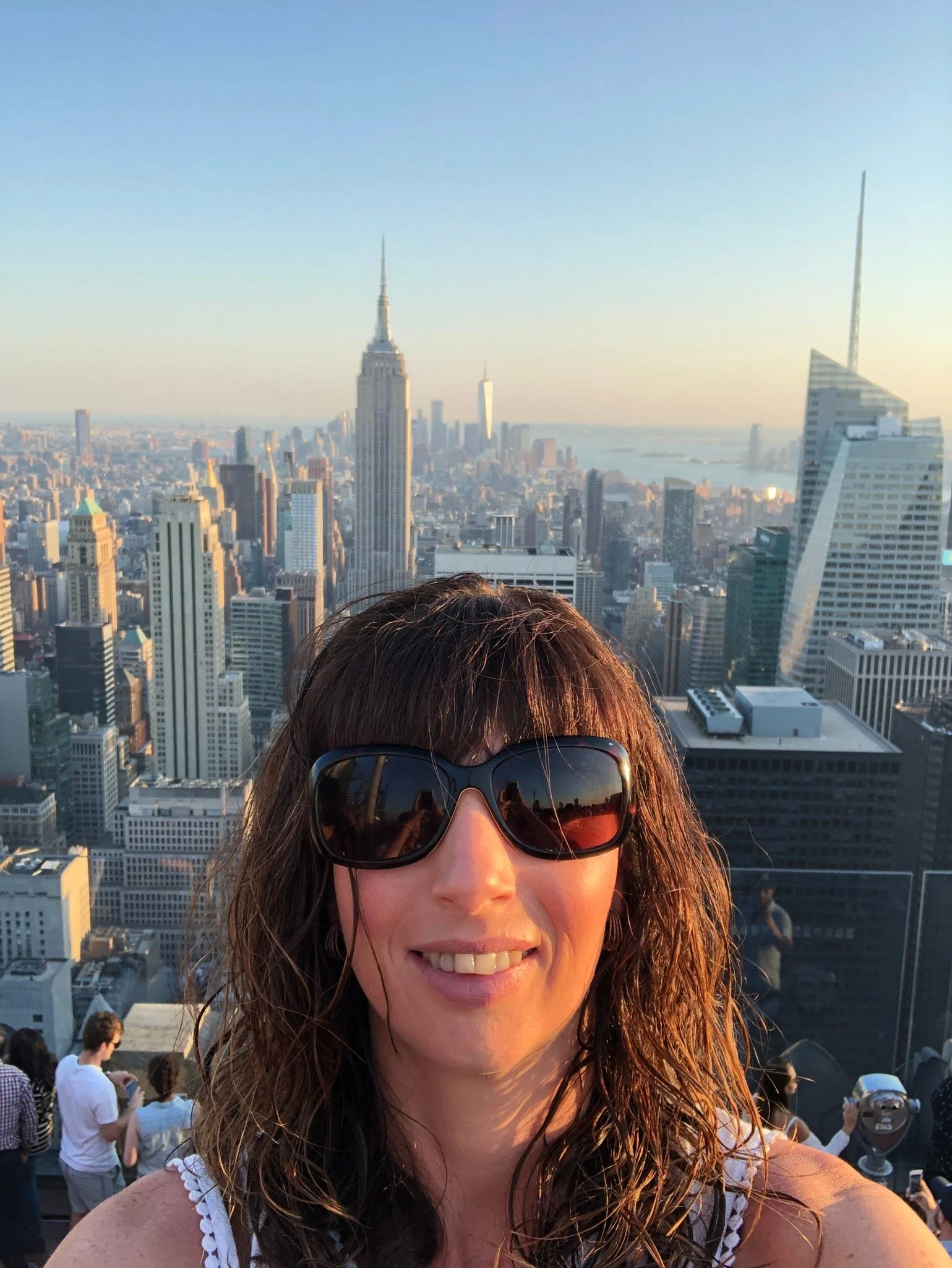 Smiling woman with sunglasses taking a selfie on a rooftop with New York City skyline in the background.