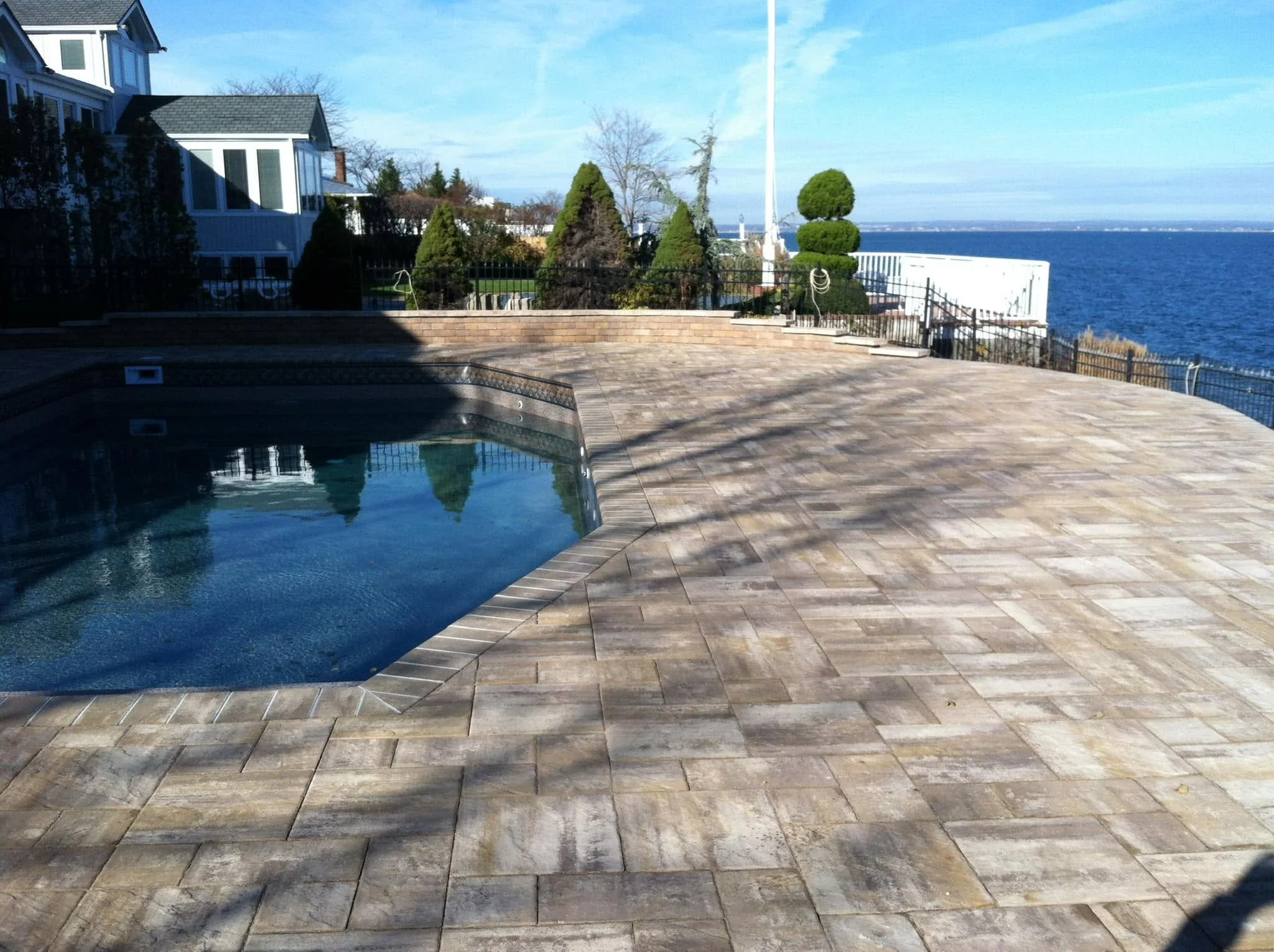 View of a backyard with a hot tub, stone patio, landscaped trees, and a view of the ocean in the distance.