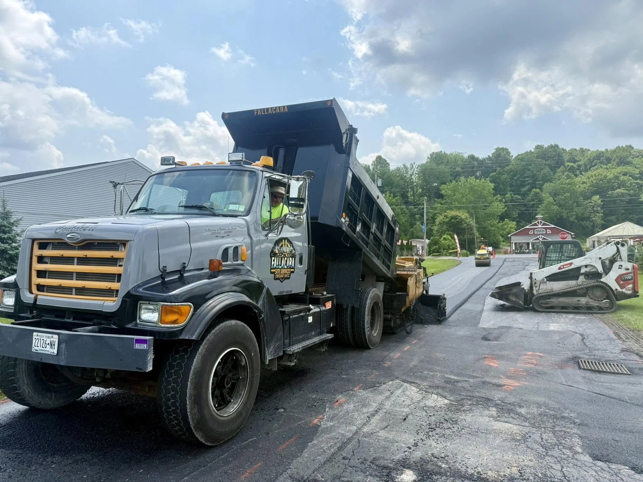 A large dump truck with the label 'Fallacara' on the back, pouring asphalt onto a road under construction, with construction equipment nearby and a small building and trees in the background.