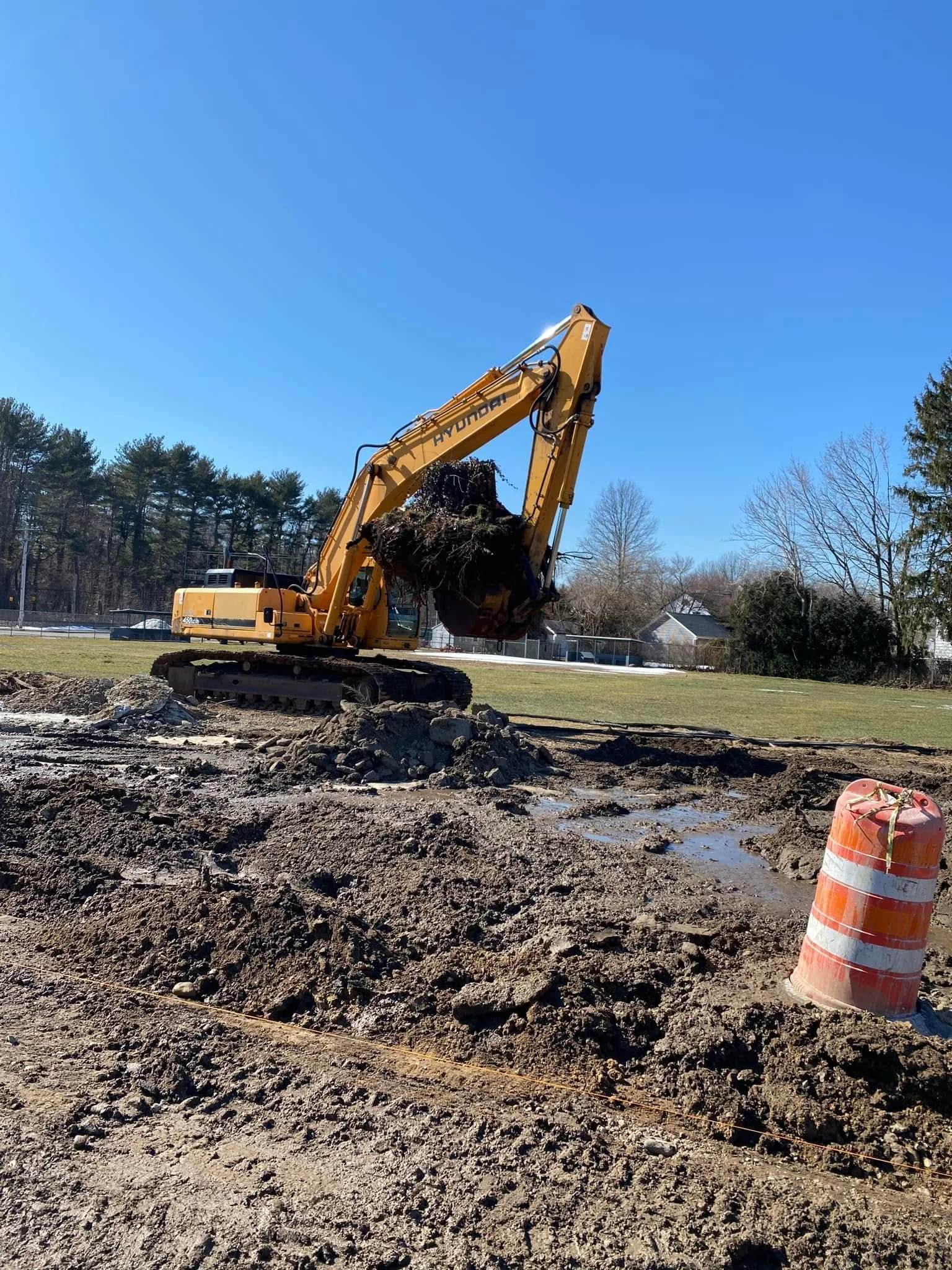 Construction site with a yellow excavator lifting dirt, orange traffic barrel in foreground, muddy ground with disturbed soil, trees and houses in background, clear blue sky.