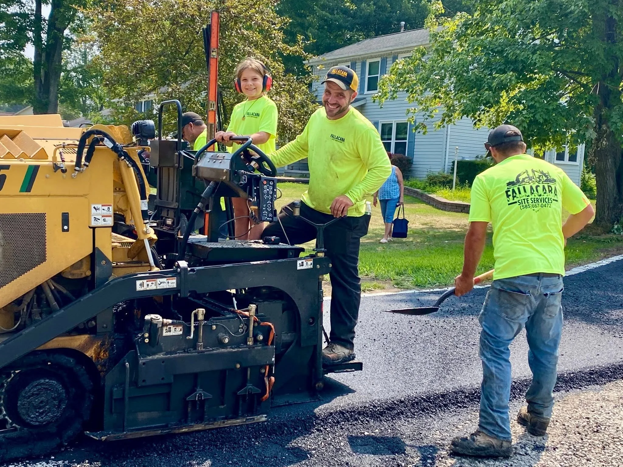 Workers paving a residential street, with a young girl smiling and wearing ear protection, and a man operating a paving machine surrounded by green trees and houses.
