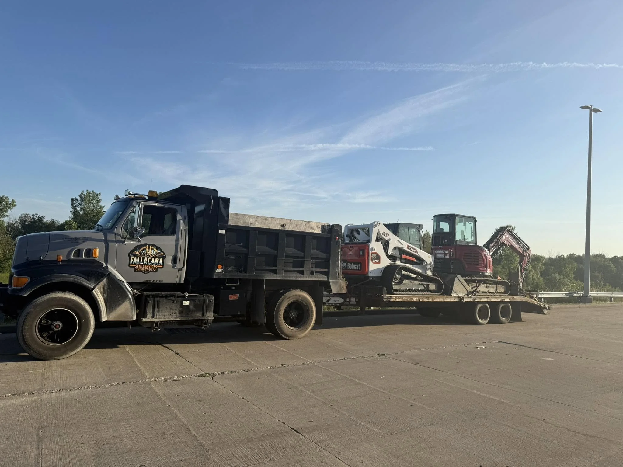 A flatbed truck transporting two compact excavators parked on the side of a paved road with trees and a clear sky in the background.