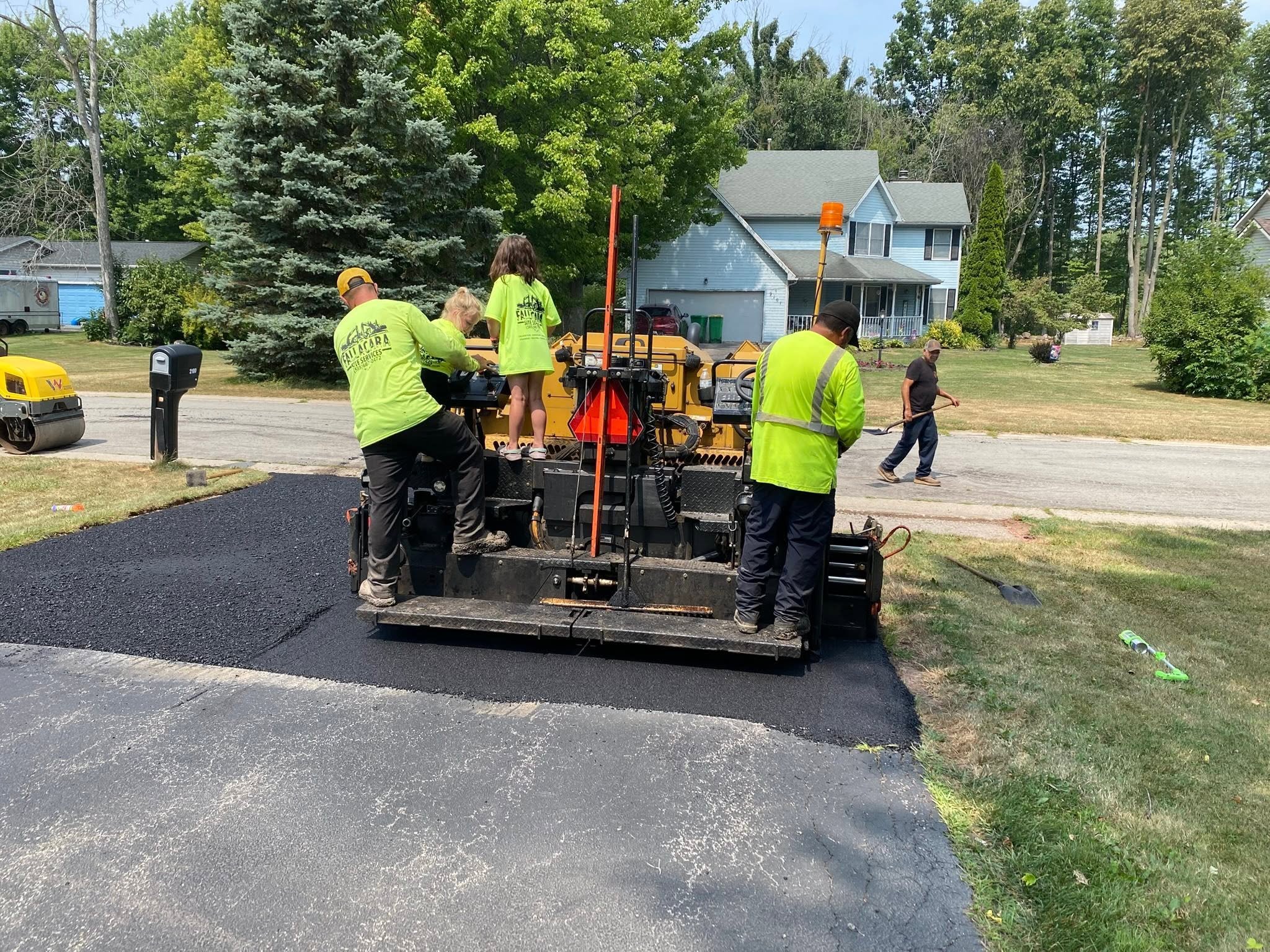 Workers in yellow and green shirts are paving a street with fresh asphalt, with a blue house and trees in the background.