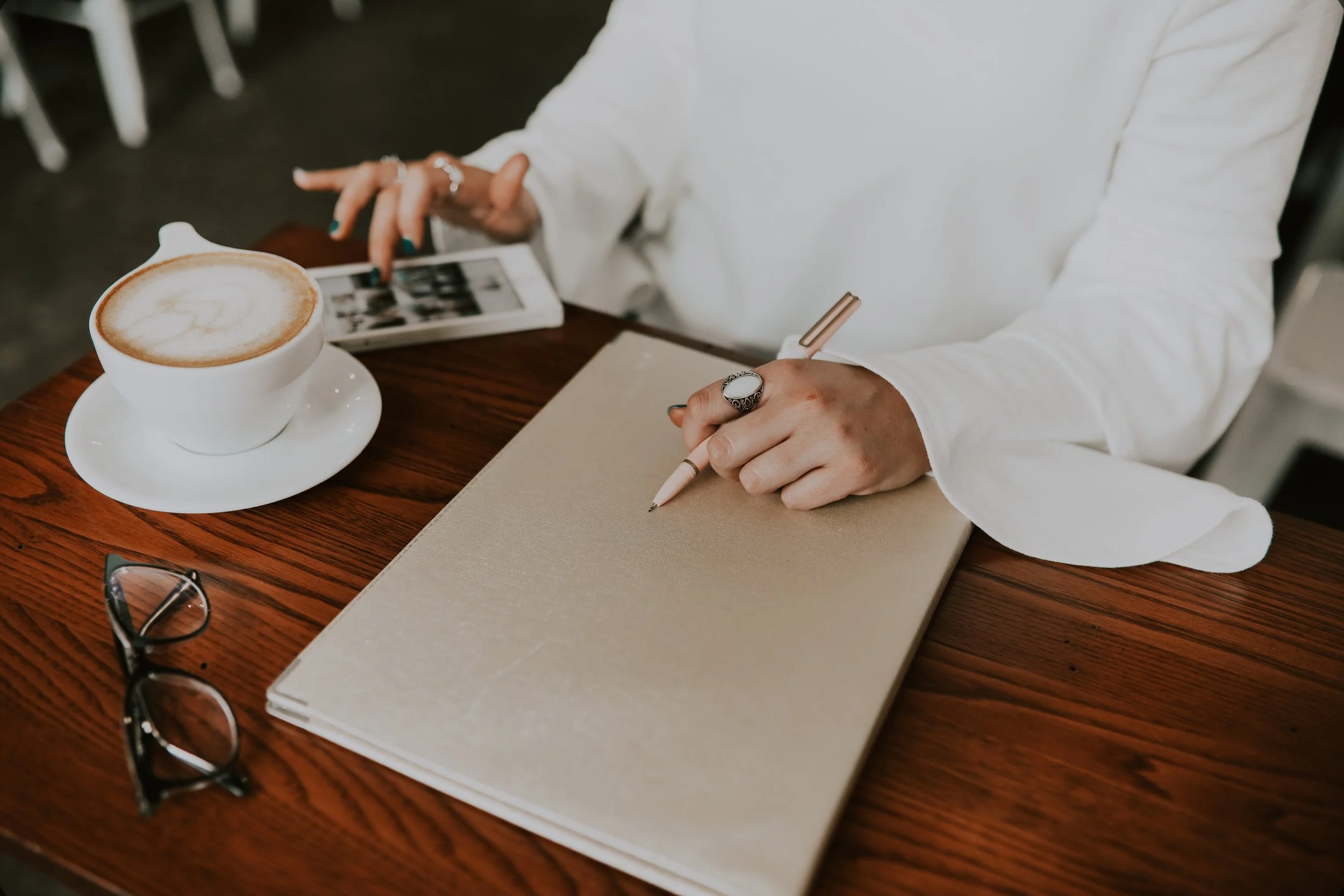 Person wearing white shirt using a tablet and holding a pen, with a cup of coffee on a wooden table, a notebook, and a pair of glasses beside.
