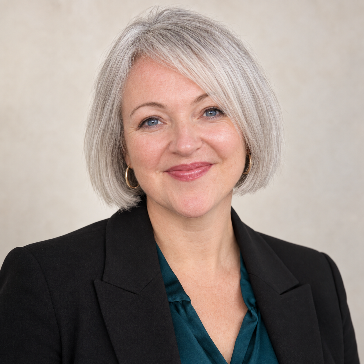 A portrait of a woman with short gray hair, blue eyes, and a warm smile. She is wearing a black blazer and a dark teal blouse, with a light, neutral background.