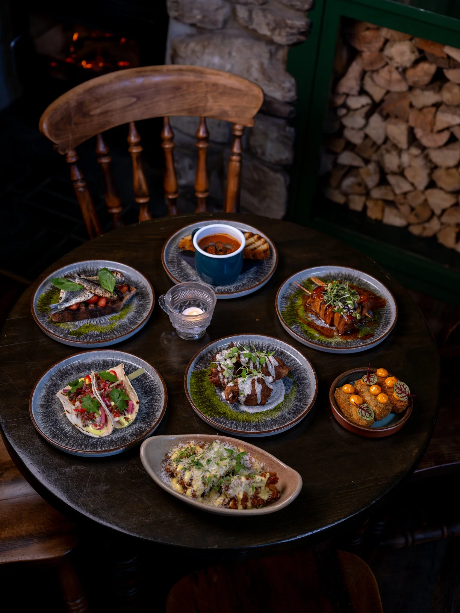 A round wooden table with various plates of gourmet food, including fish, grilled vegetables, and a small cup of soup, set in a rustic restaurant with stacked firewood and stone walls in the background.