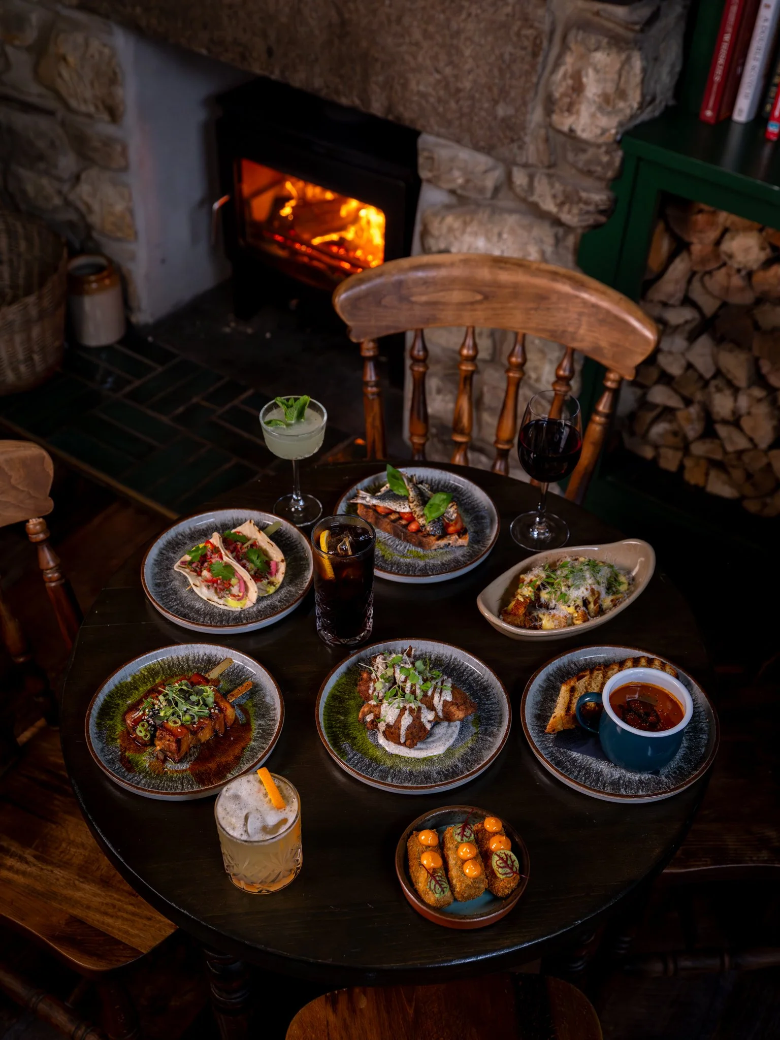 A table with various plates of gourmet food, two drinks, and a fireplace in the background.