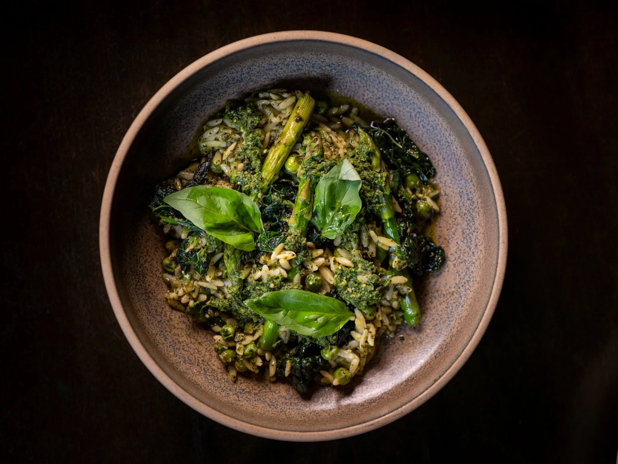 A bowl of green pasta with leafy greens, herbs, and grains on a dark surface.
