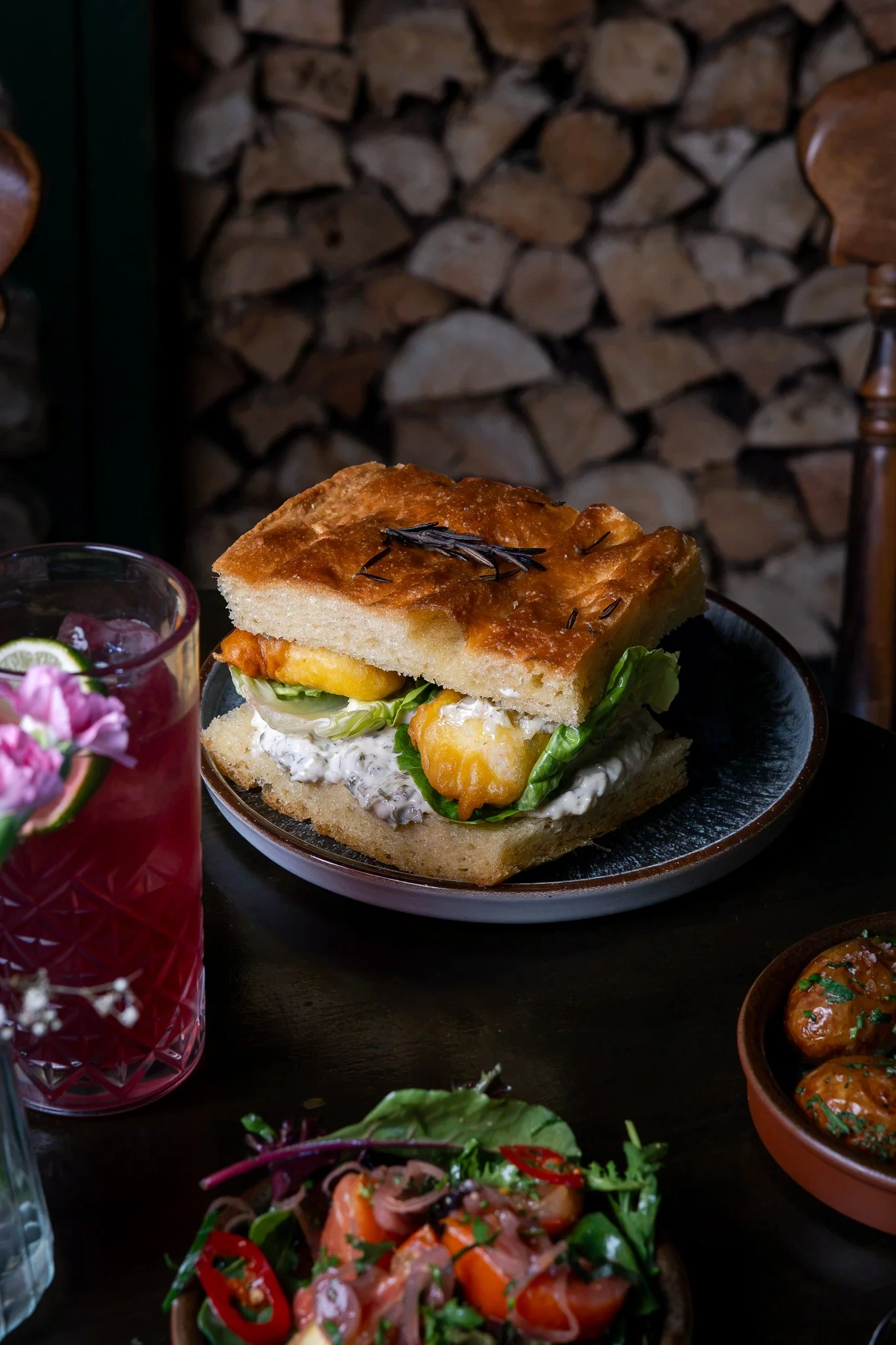 A slice of focaccia bread with herbs on top, served on a plate with lettuce and a creamy spread underneath, surrounded by salads and drinks in a rustic setting.