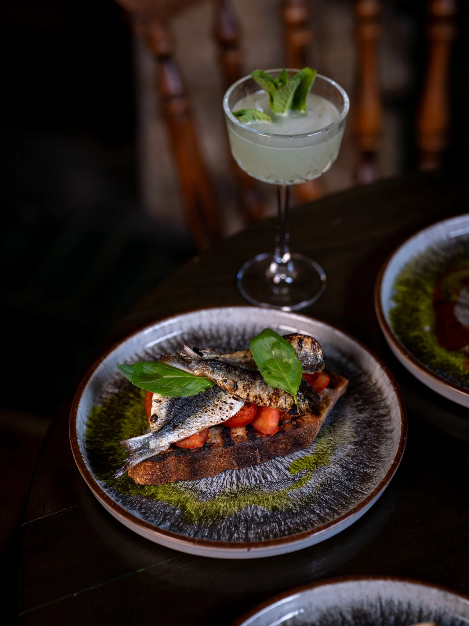 A plate with a piece of toasted bread topped with diced tomatoes and two small grilled fish garnished with fresh basil leaves, served on a patterned ceramic dish.
