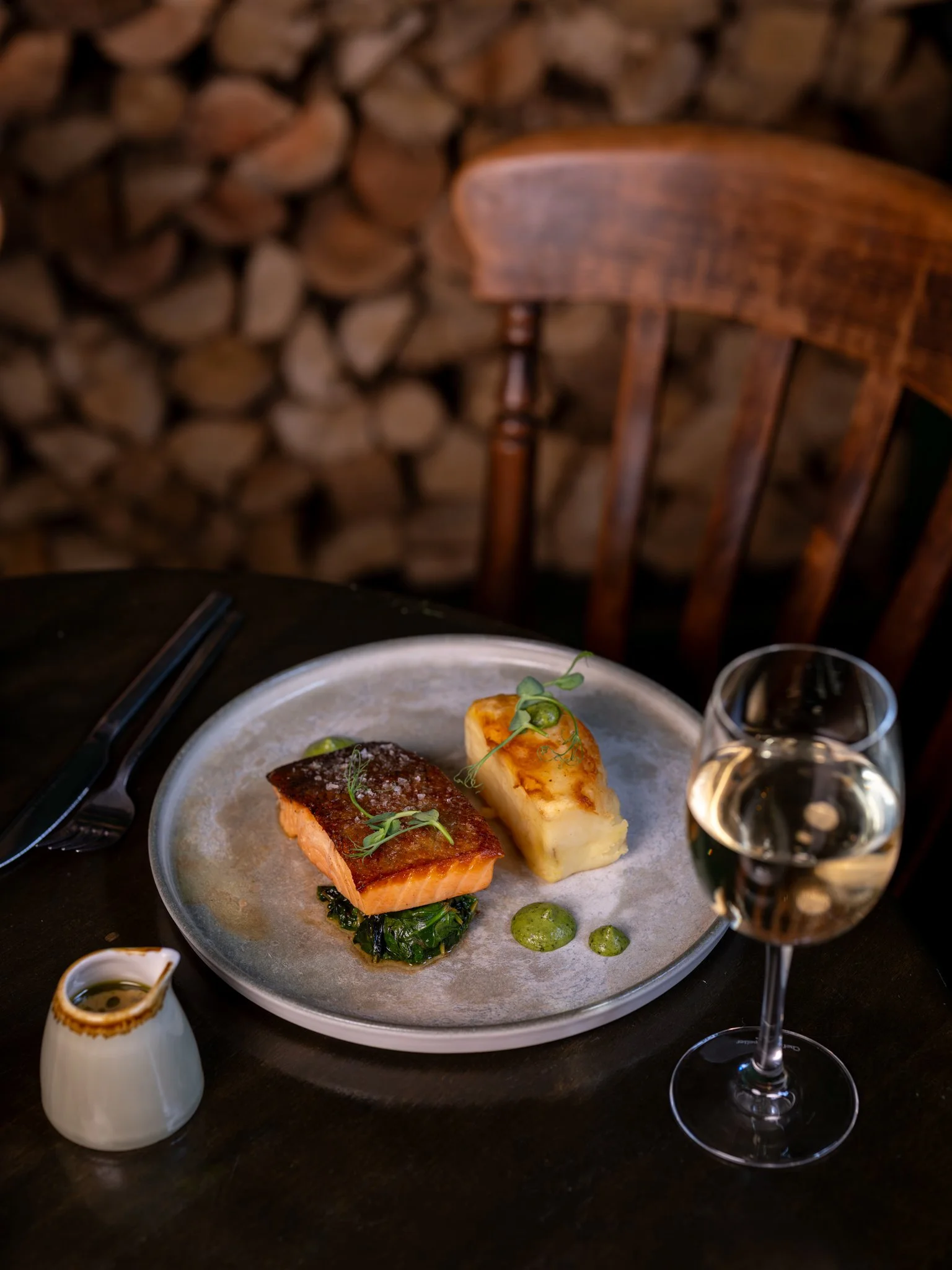A plate with cooked salmon, a piece of grilled cheese, and green sauce garnished with herbs on a dark table. There is a glass of white wine and a small pitcher with sauce nearby, with a wooden chair and stacked firewood in the background.