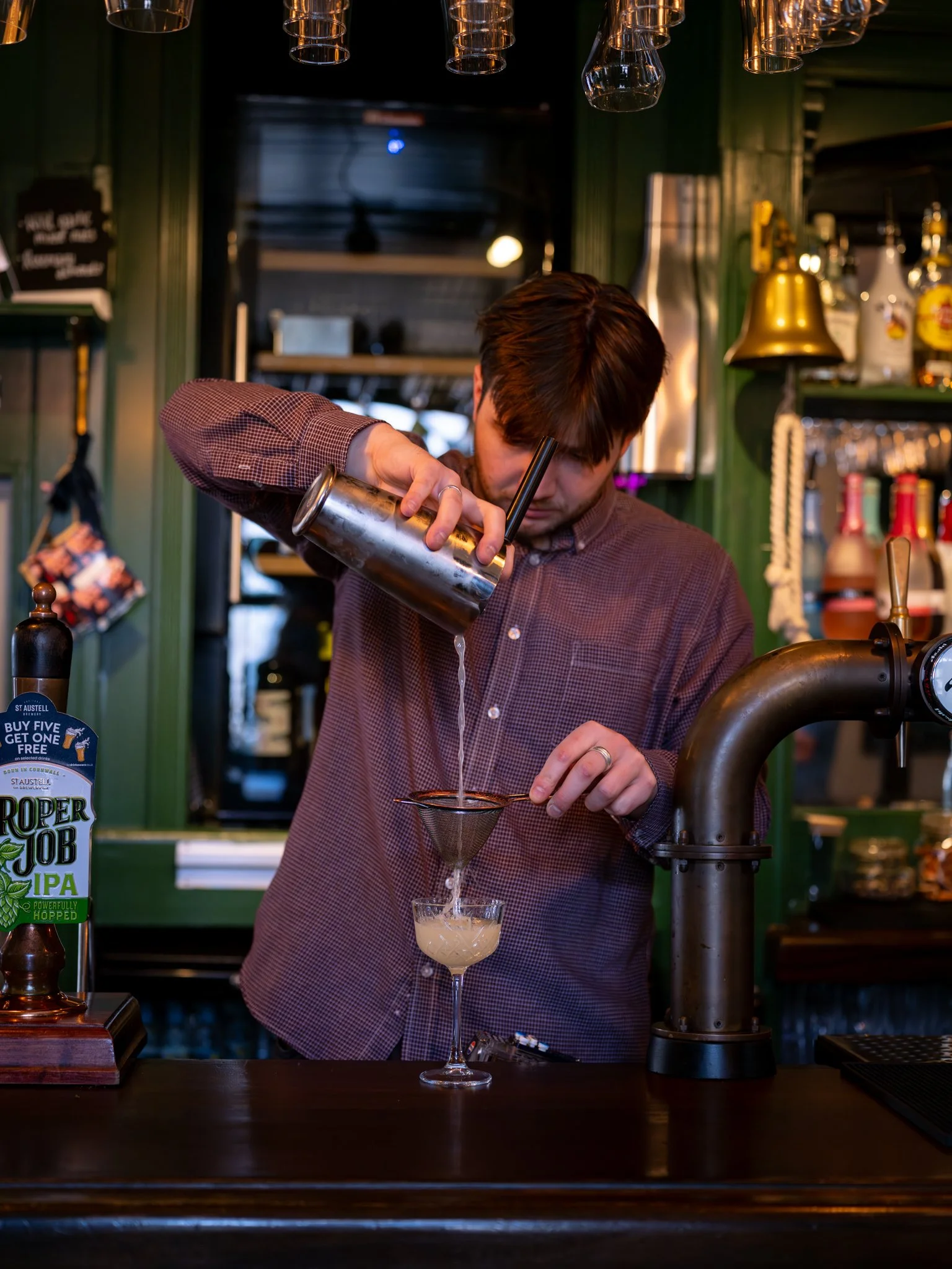 A bartender pouring a liquid through a strainer into a cocktail glass at a bar with green walls and shelves of bottles.