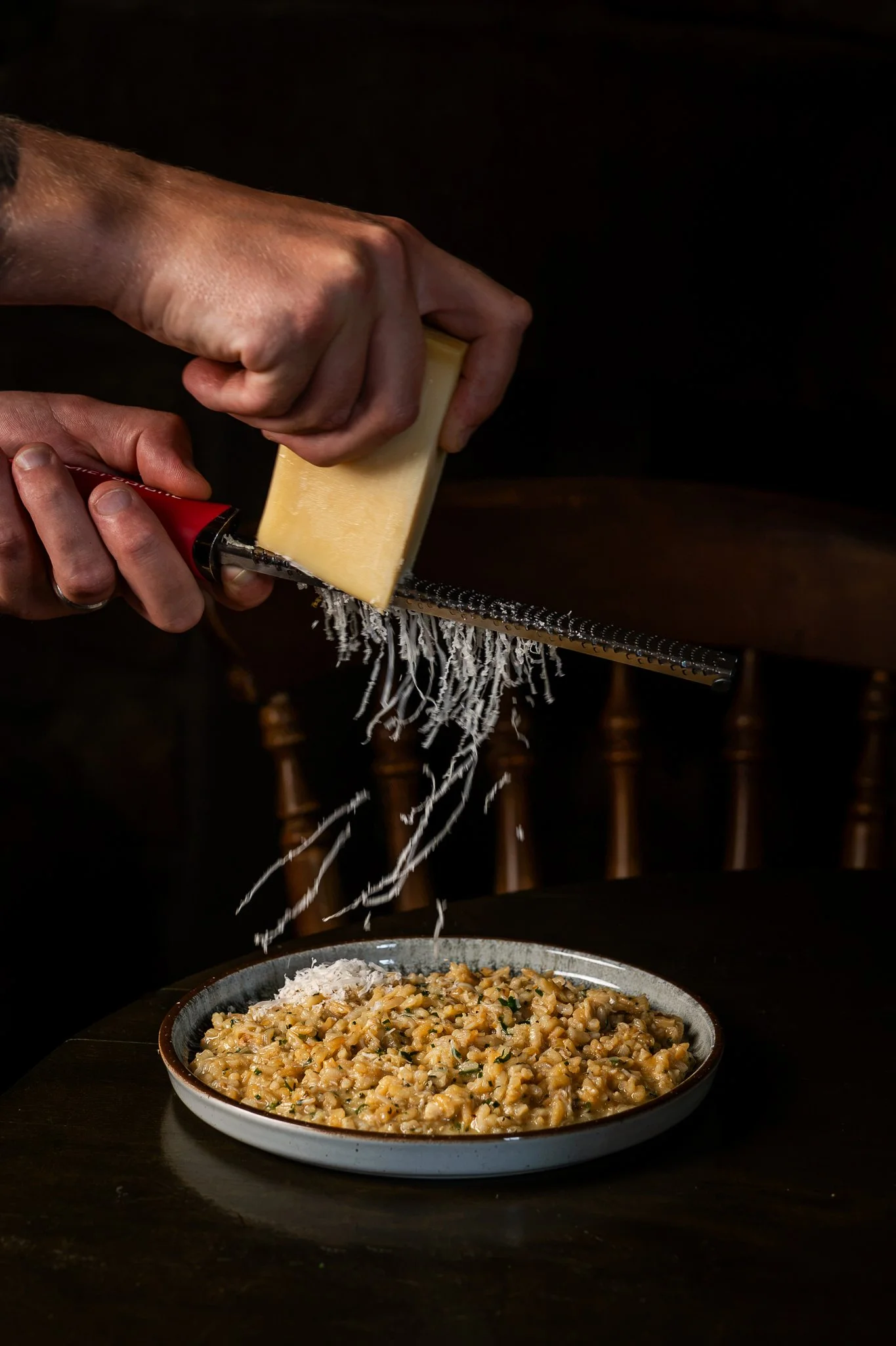 Person grating cheese over a plate of cooked risotto on a dark wooden table, with chairs in the background.