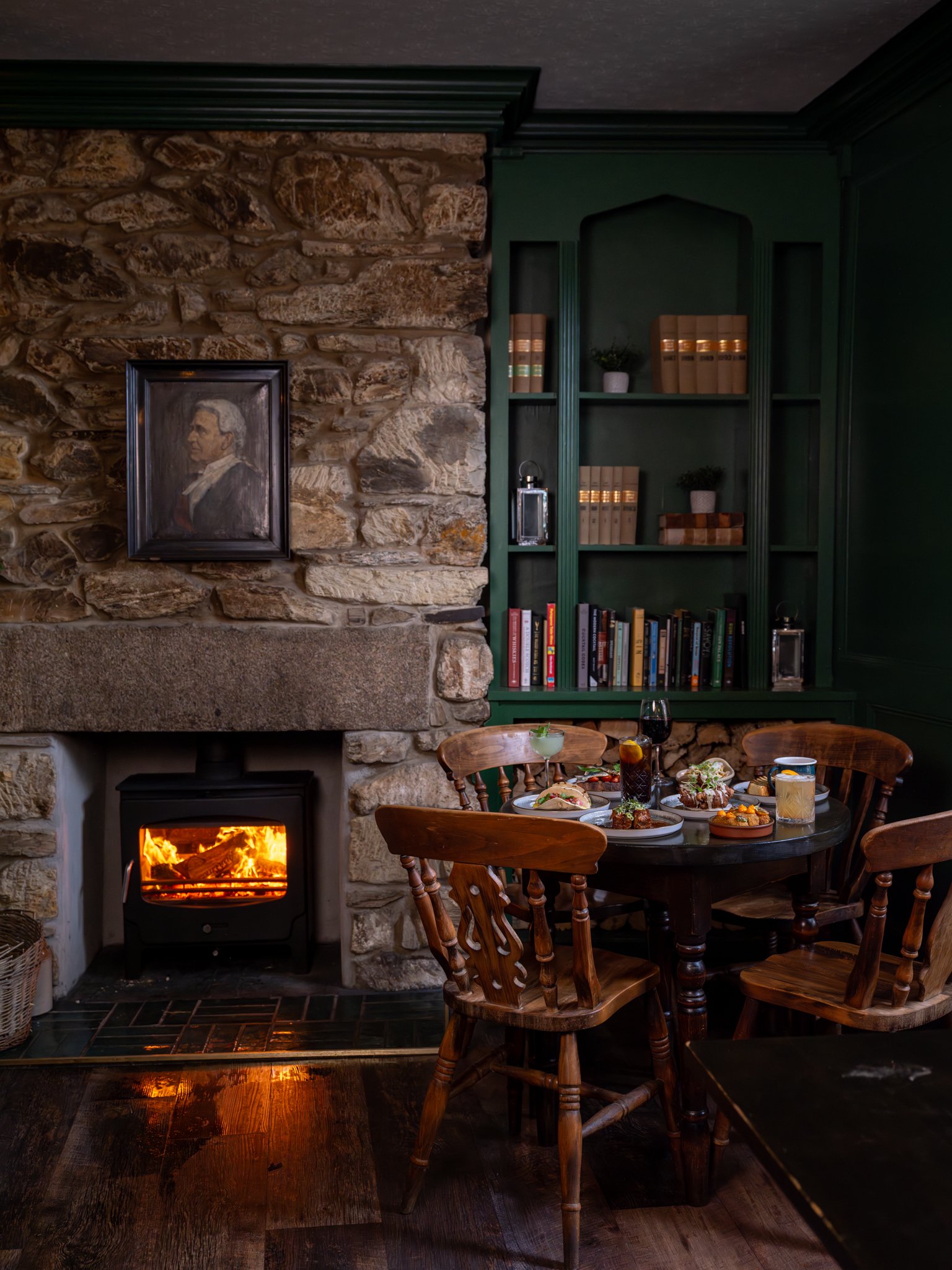 A cozy dining area with a stone fireplace lit, a dark green bookshelf filled with books and decorative items, a round wooden table with chairs, and various dishes and drinks set for a meal.