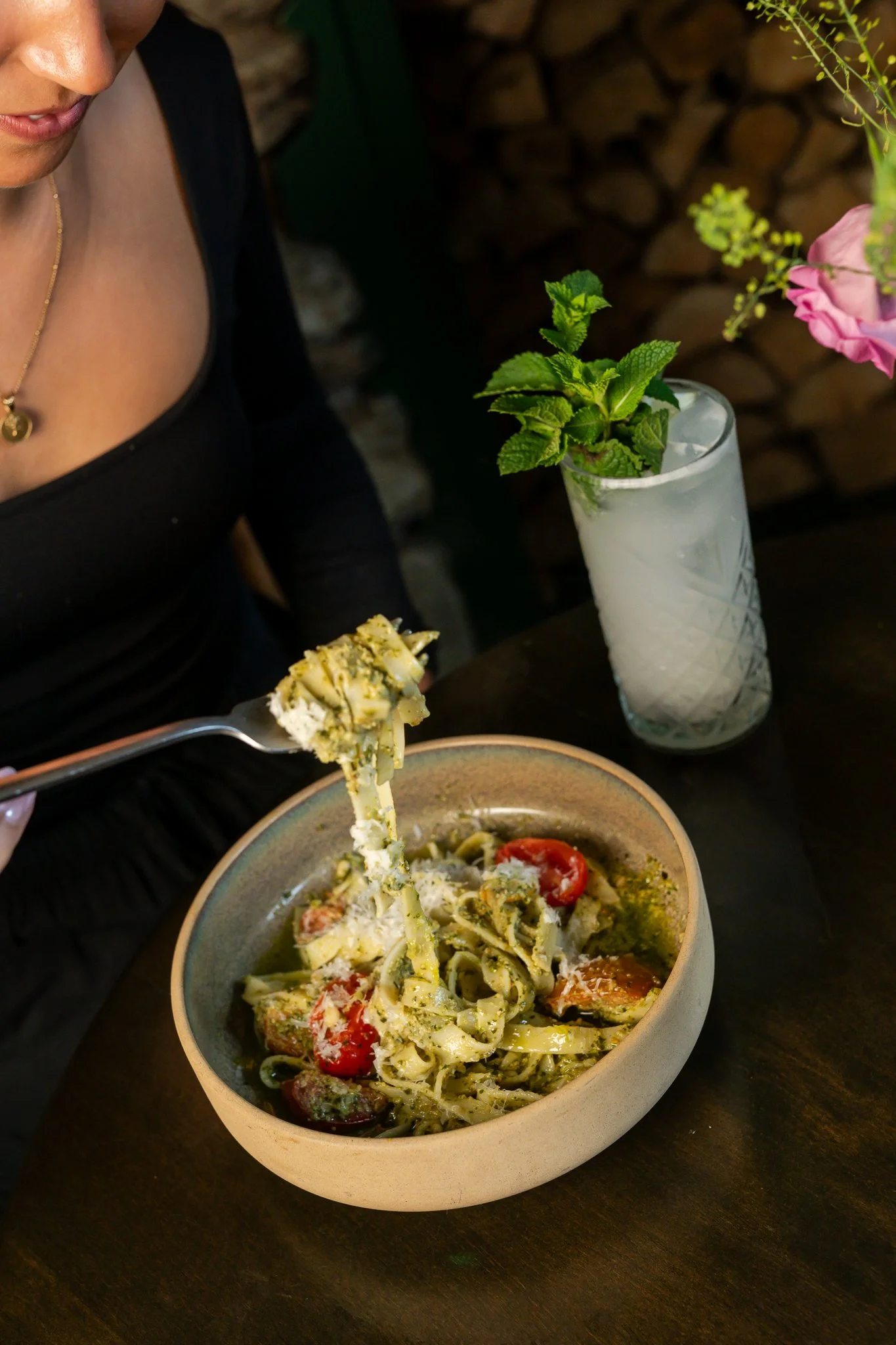 A bowl of pasta with cherry tomatoes and herbs, and a fork lifting a portion. A woman's face partially visible, sitting at a dark table with a vase of flowers and a drink.