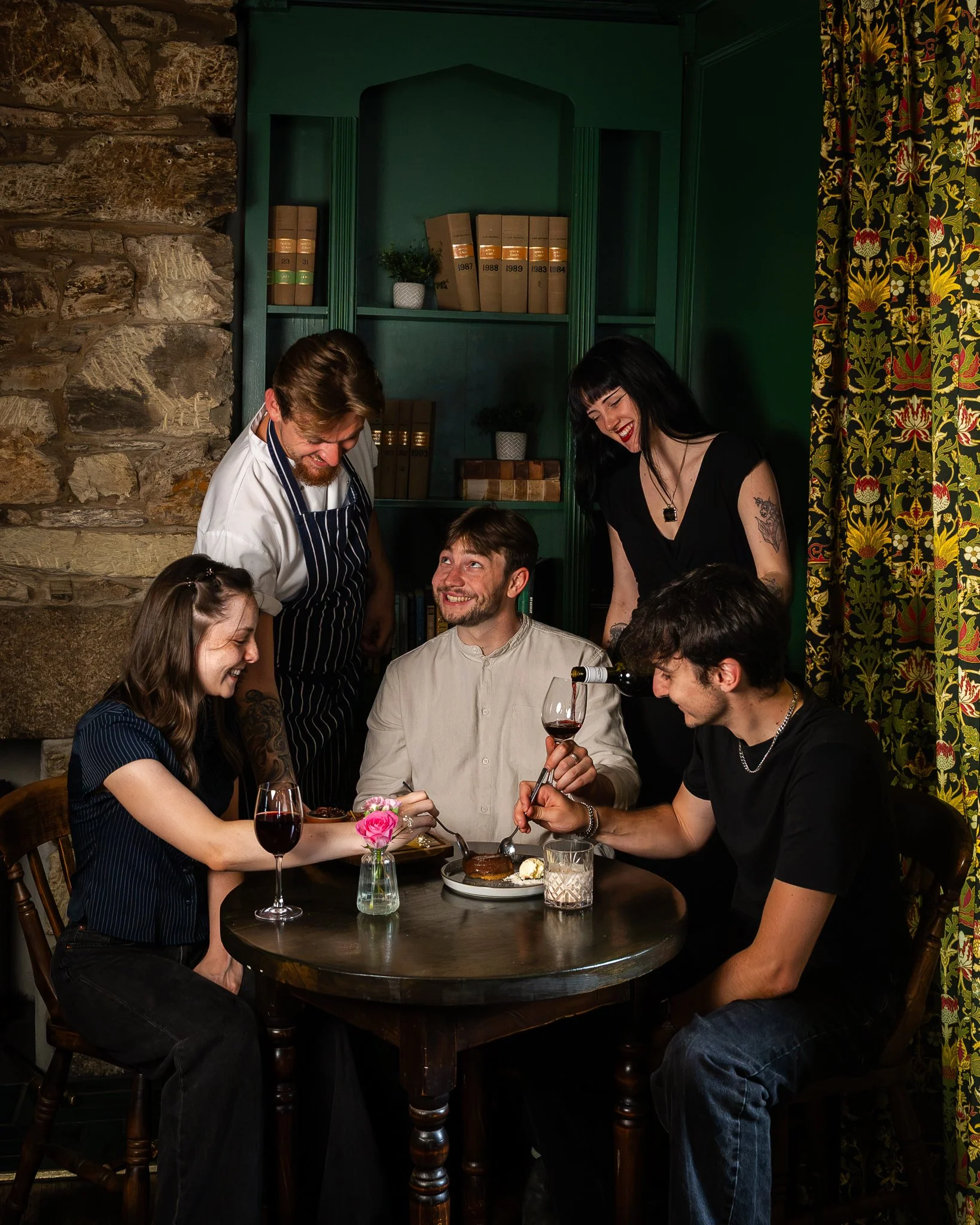 A group of people gathered around a table in a cozy restaurant, smiling and enjoying each other's company while sharing a meal. A woman is serving food, and a man is pouring red wine. The background features a green bookshelf with books and decorative items, stone wall, and colorful patterned curtains.