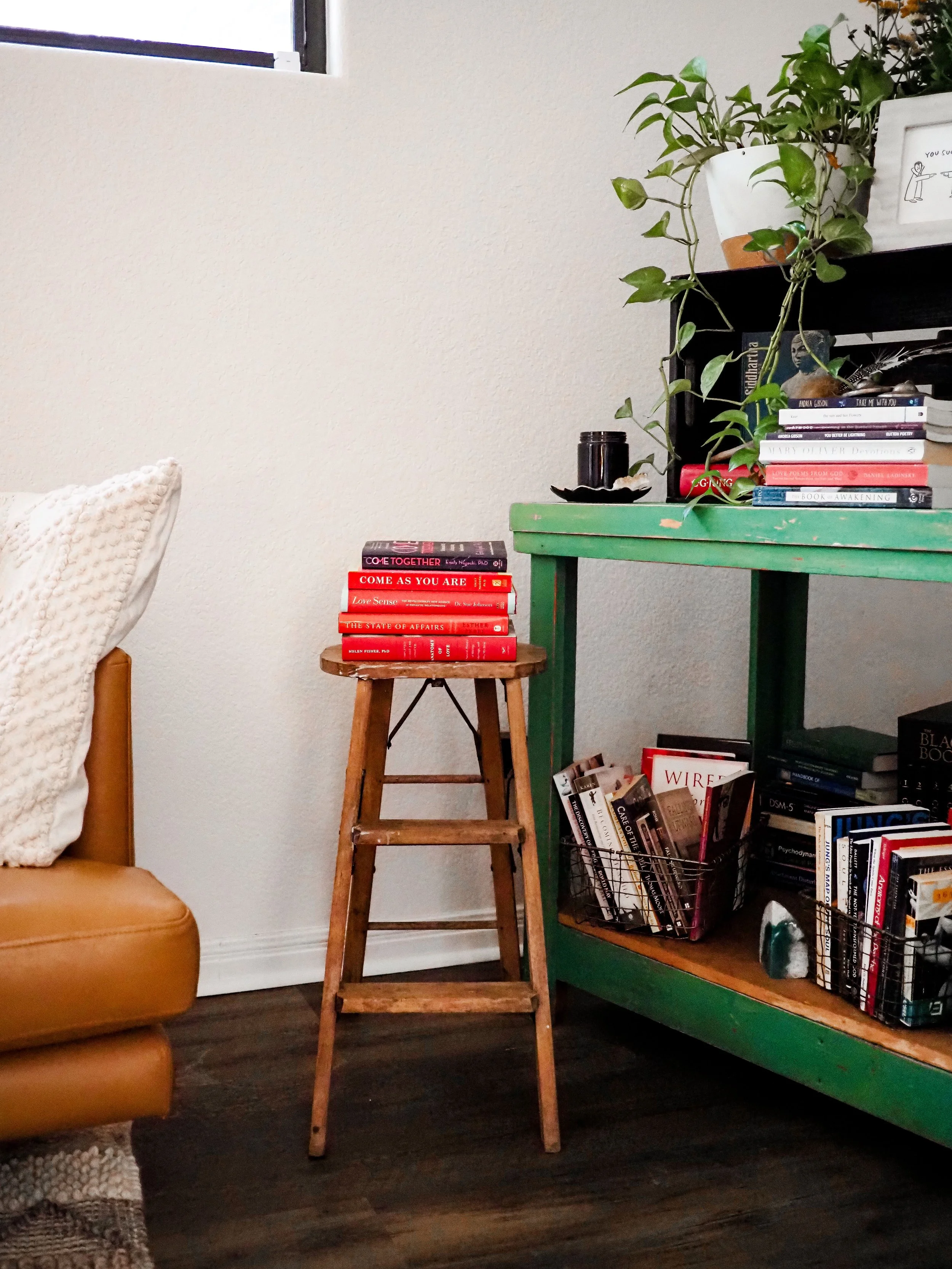 Cozy living room with a leather chair, wooden step stool, green console table, houseplants, and stacks of books.