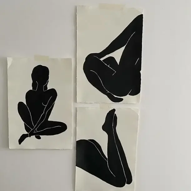 Three black and white silhouette art pieces of human figures on white paper, arranged on a wall. The artwork depicts a person sitting cross-legged, a crouching pose, and a person with one lifted leg.