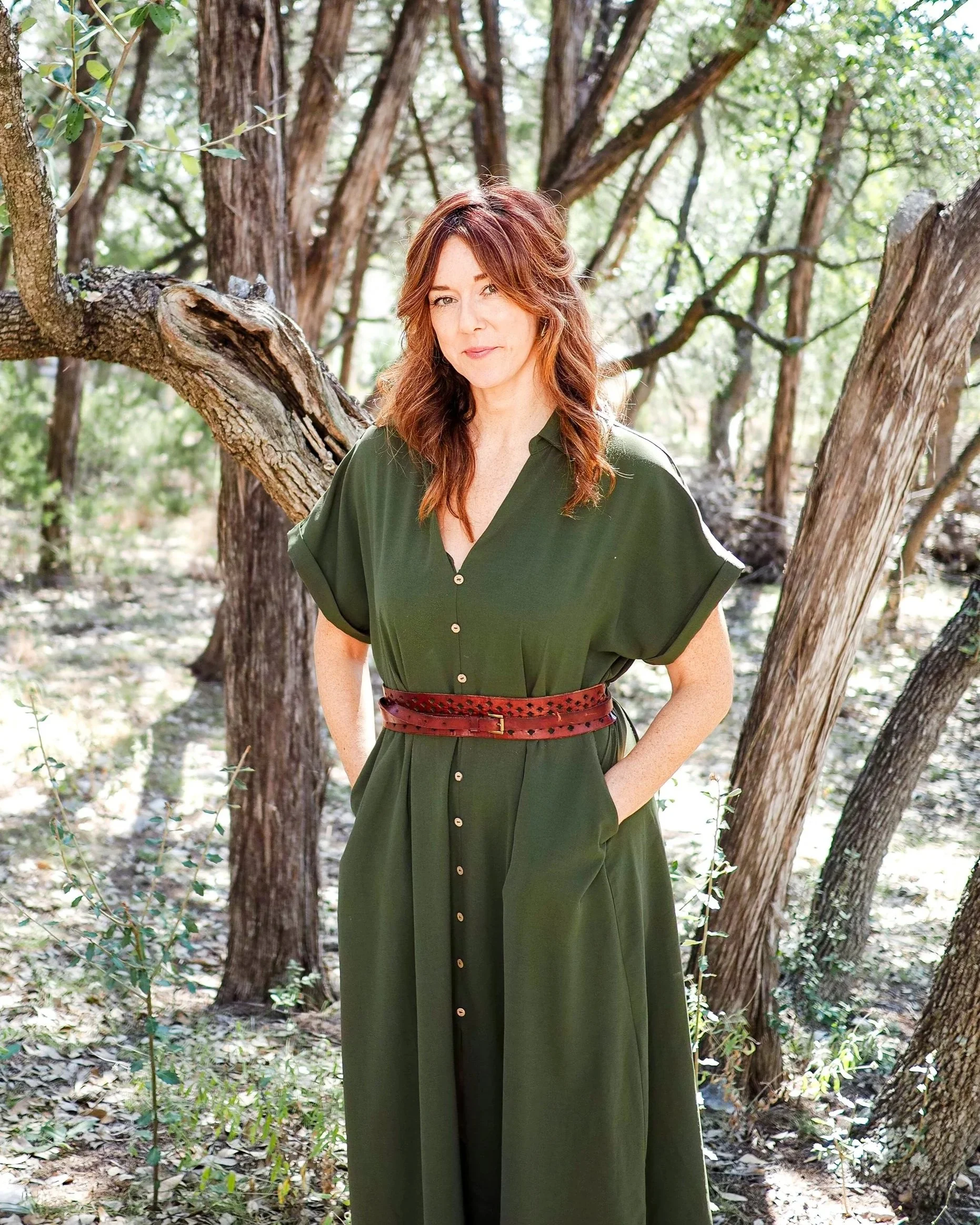 Woman with auburn hair wearing a green dress and red belt standing in a sunlit forest