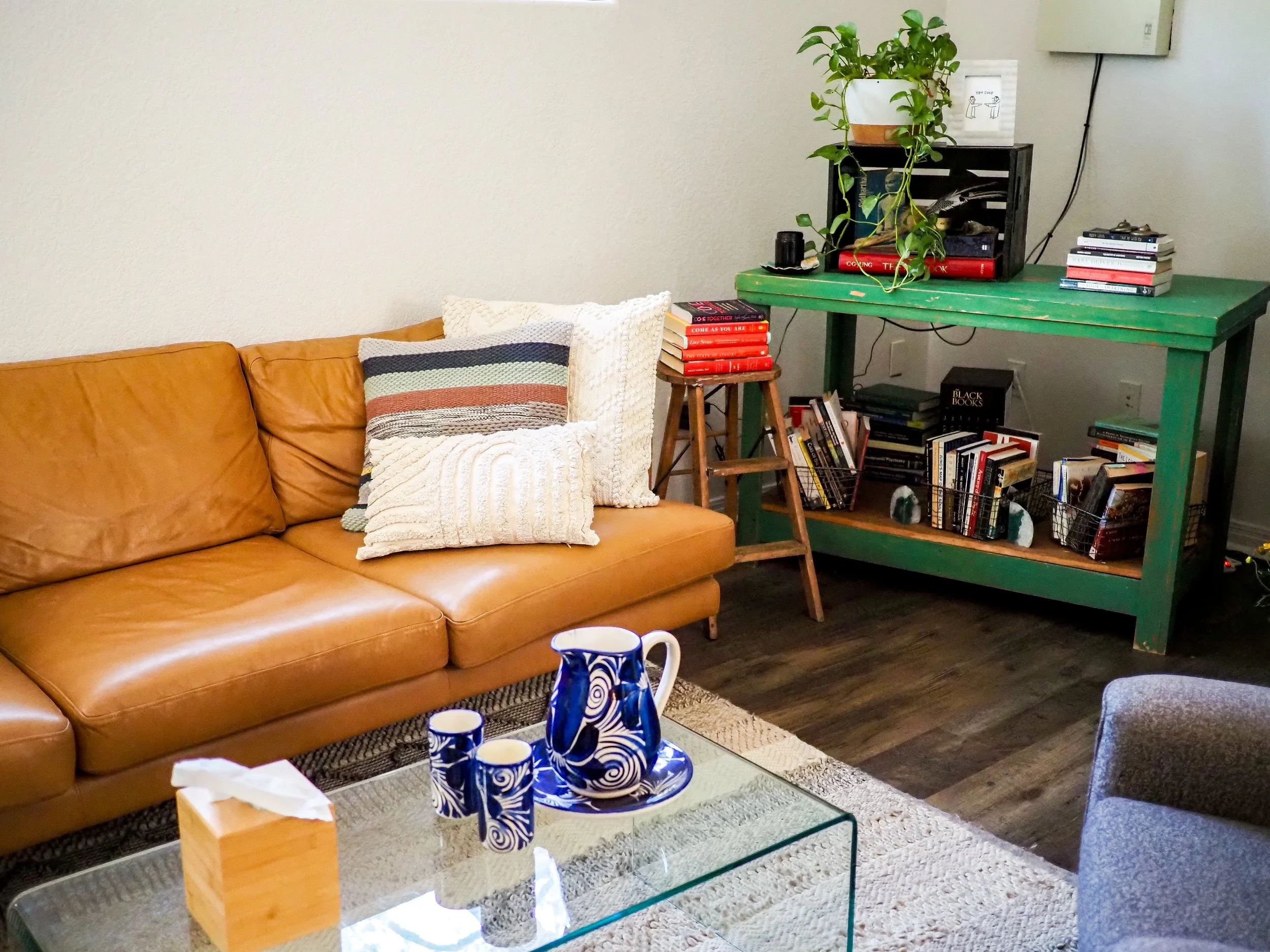 Cozy living room with a caramel leather sectional, patterned pillows, glass coffee table, blue-and-white pitcher set, green bookshelf, books, and potted plant