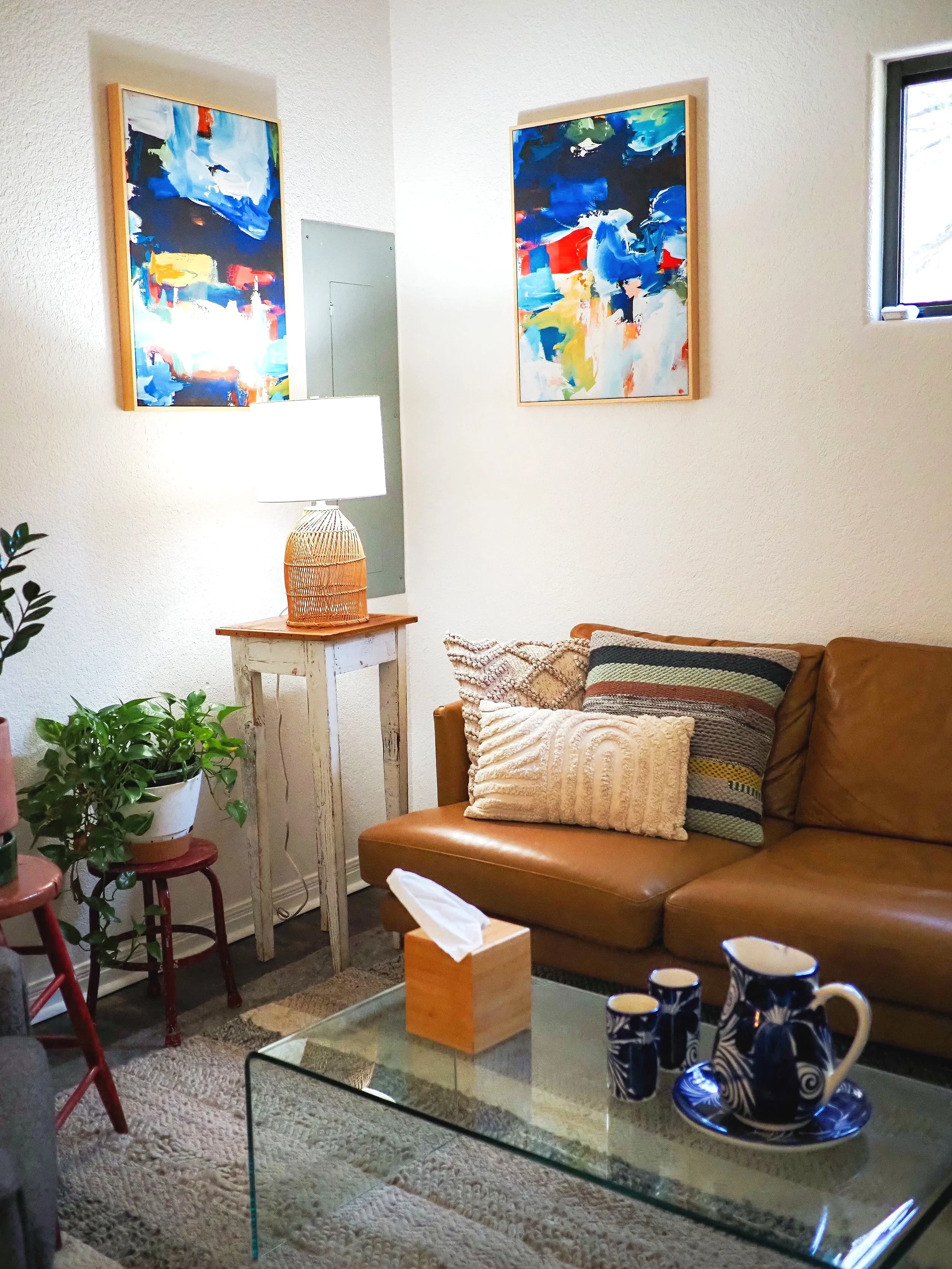 Cozy living room with colorful abstract paintings, leather sectional sofa, woven lamp, houseplant, and glass coffee table.