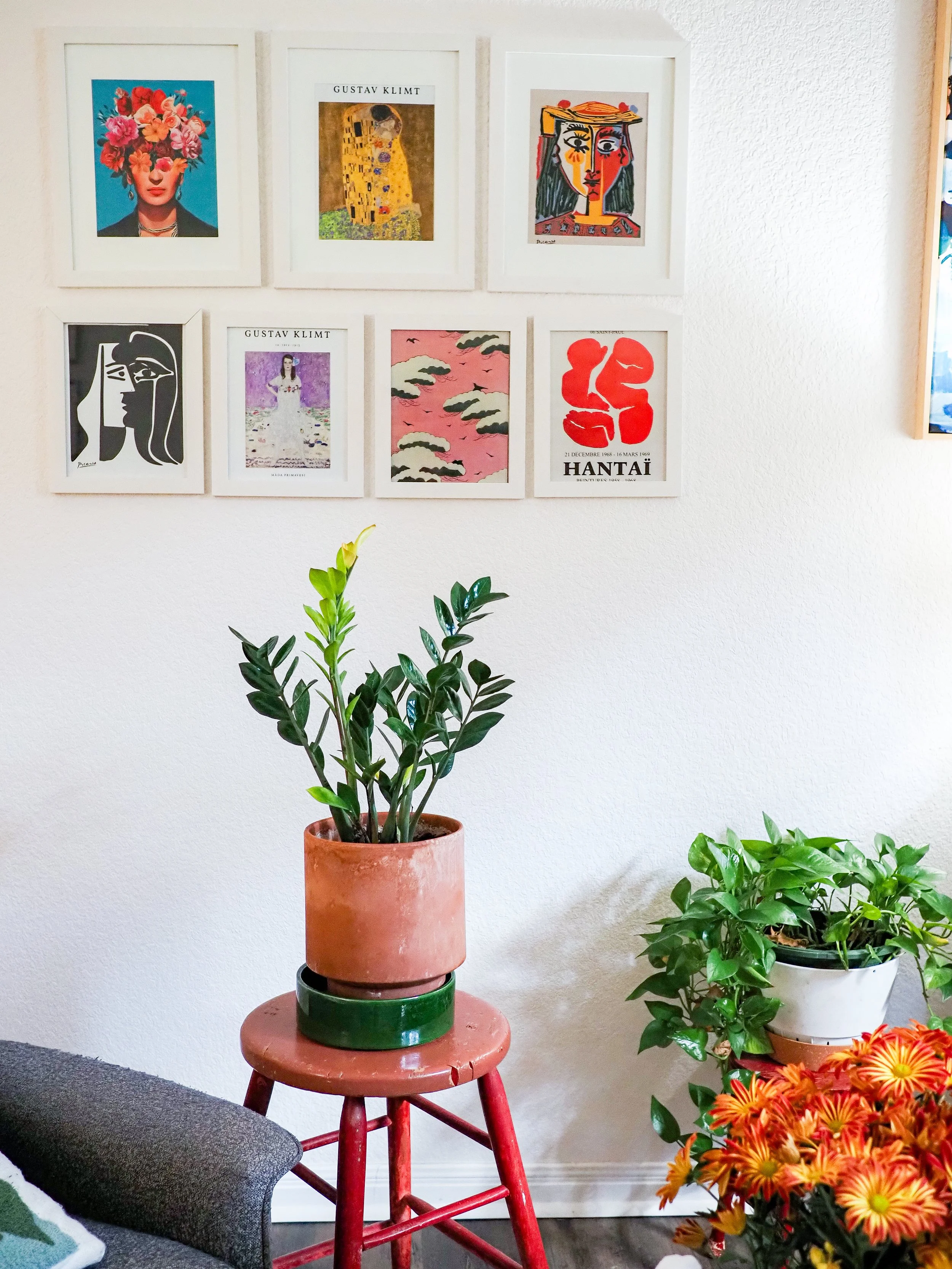 Gallery wall with colorful framed art above potted houseplants, orange flowers, and a red plant stand in a cozy living room.