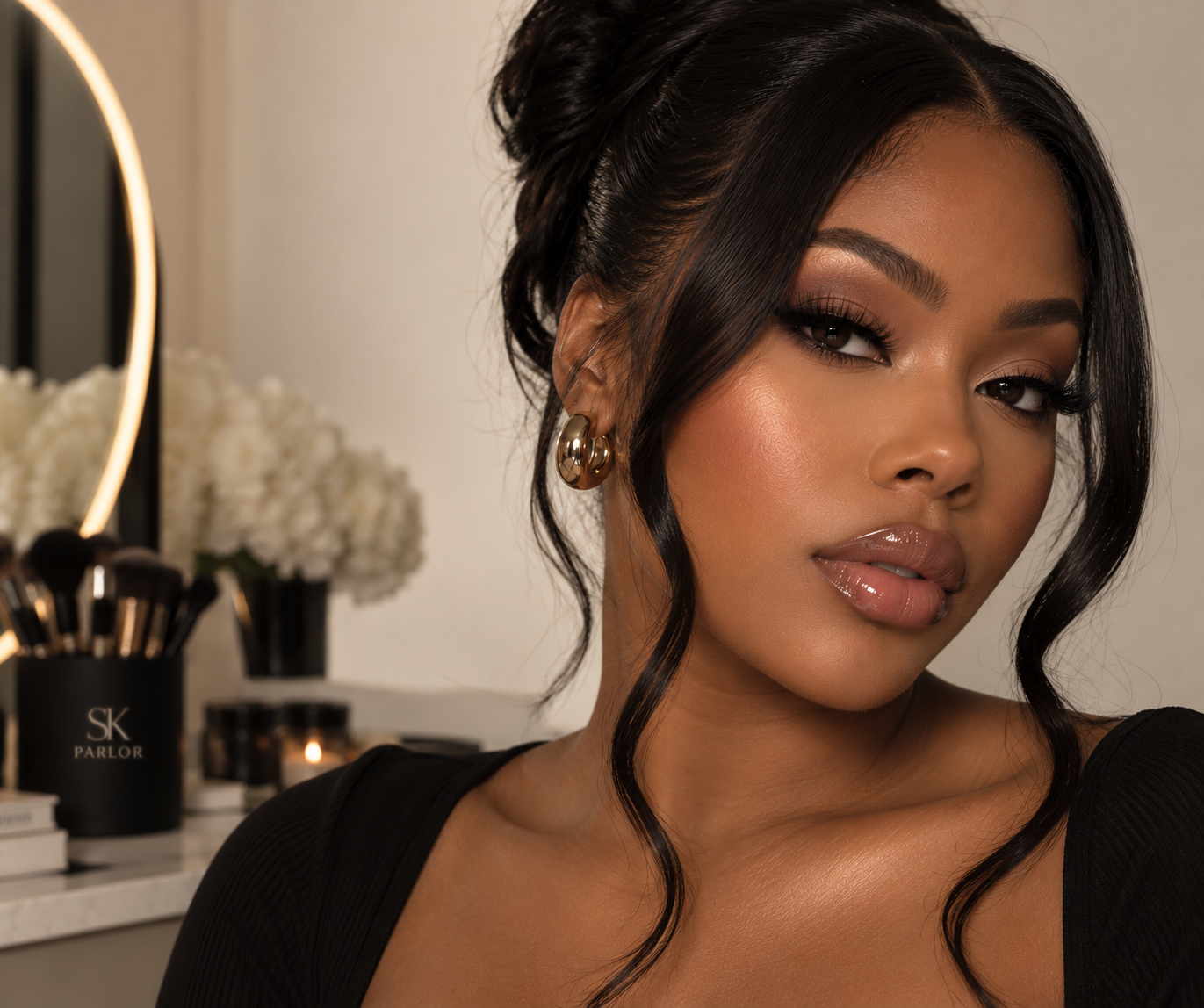 Close-up portrait of a woman with dark hair styled in loose waves, wearing makeup and gold hoop earrings, in a room with a mirror, makeup brushes, and flowers in the background.