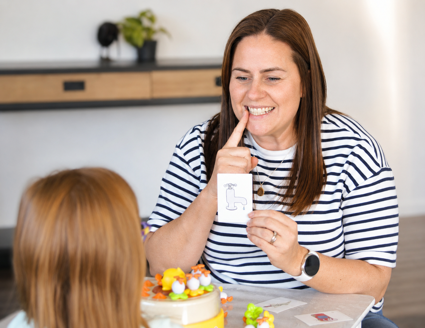 Woman with brown hair in a striped shirt smiling and holding a card with a drawing of a faucet, teaching a young girl with red hair about the faucet, on a table with a decorated cake and toy chicks.