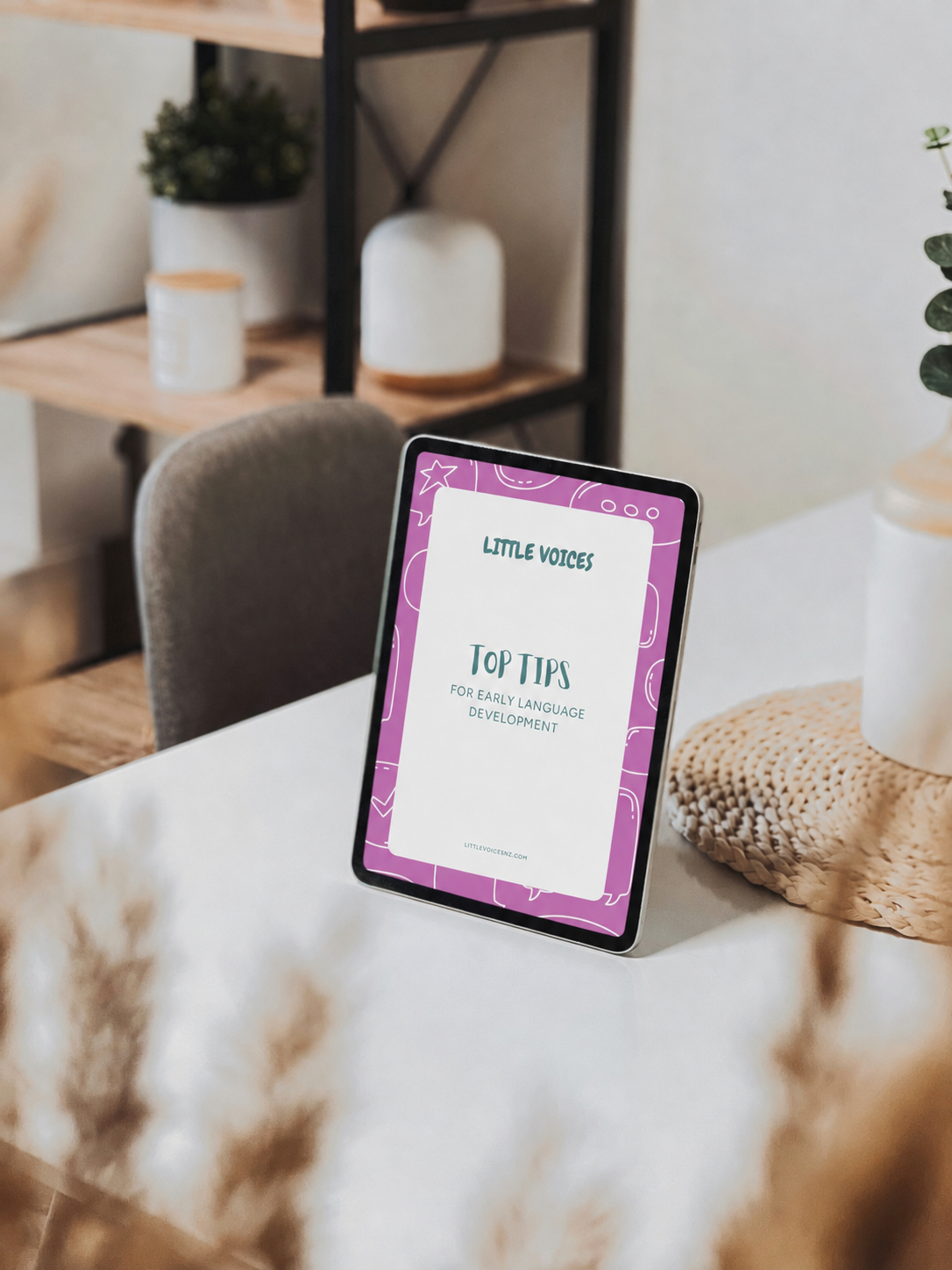 An iPad displaying a guide titled 'Little Voices: Top Tips for Early Language Development' on a white table. In the background, there is a gray chair, a kitchen with wooden shelves, and decorative vases and plants.