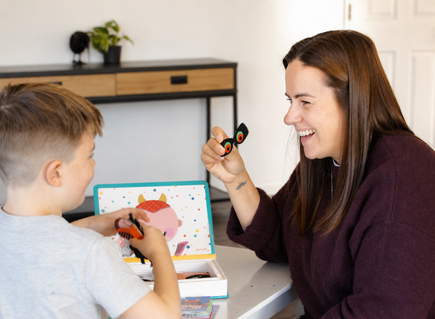 A woman and a boy smiling and playing with toy bugs at a table with a colorful photo card in the background.