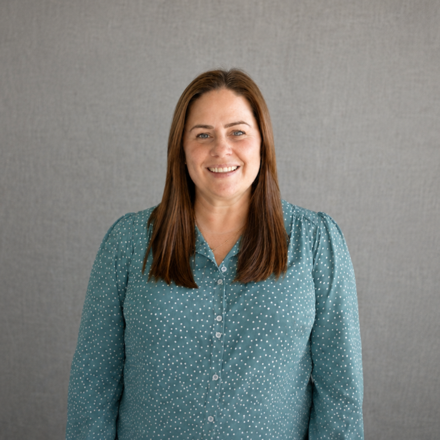 A woman with long brown hair wearing a teal blouse with white polka dots, smiling in front of a gray textured background.