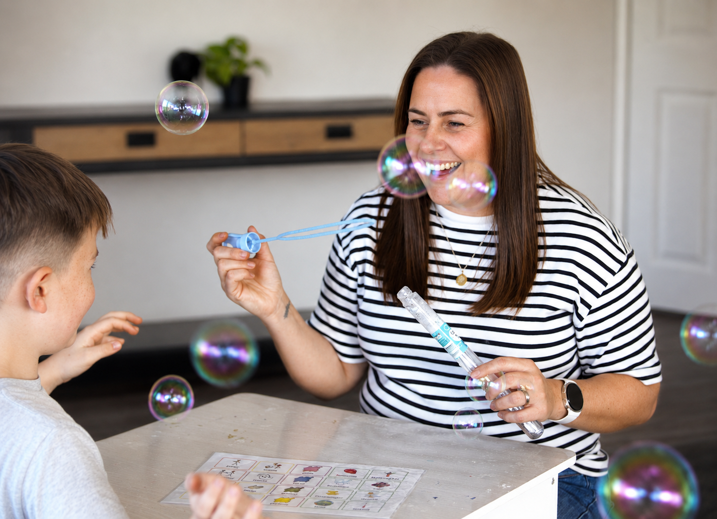 Woman blowing bubbles for a boy at a table with a bubble wand and solution, with bubbles floating around in a home setting.
