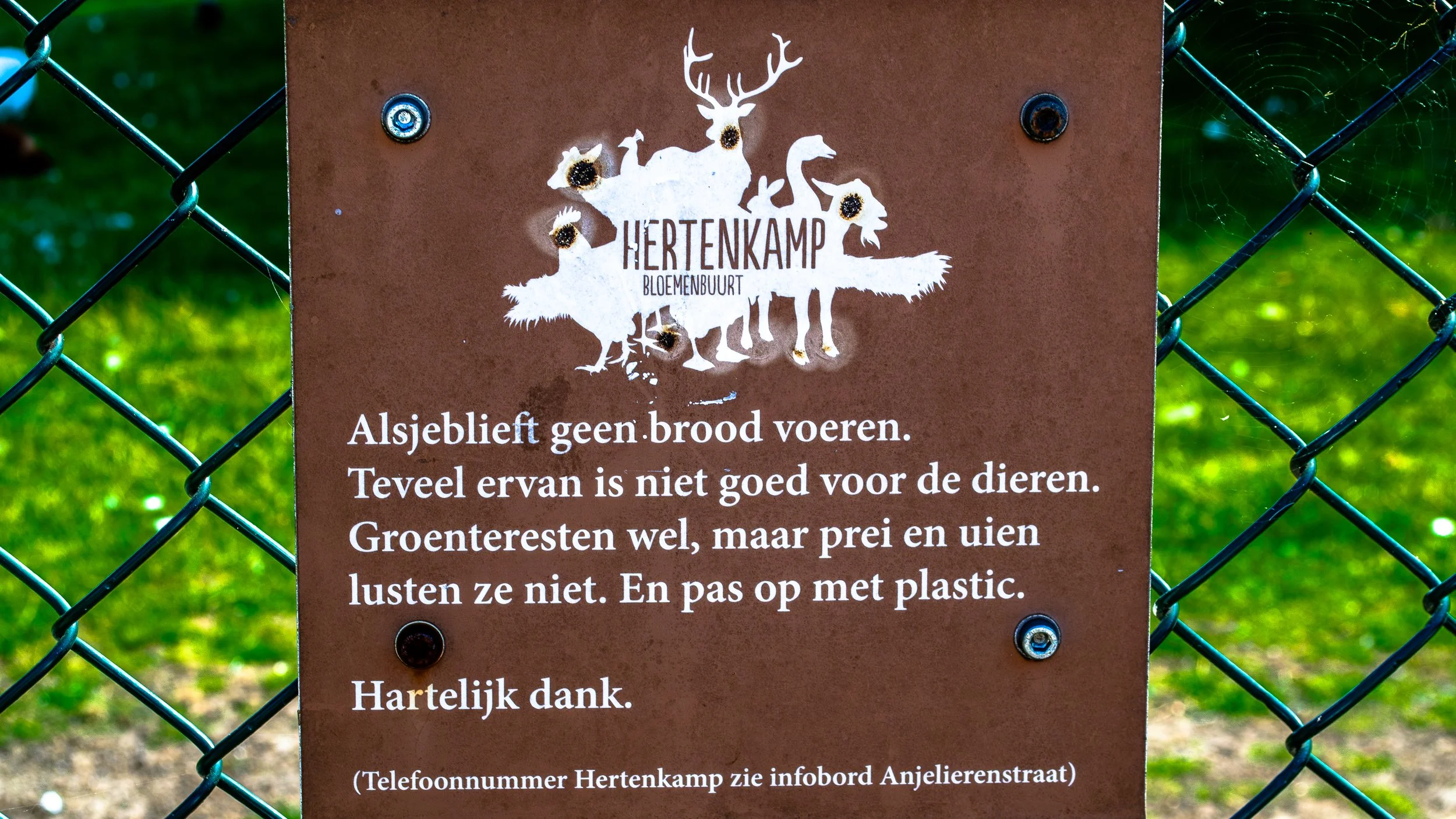A brown metal sign attached to a green chain-link fence with a spider web on the right side. The sign has a white silhouette of a fox with a deer head and antlers, and the text "Hertenkamp Bloemenbuurt" underneath. Below, there is a warning in Dutch 