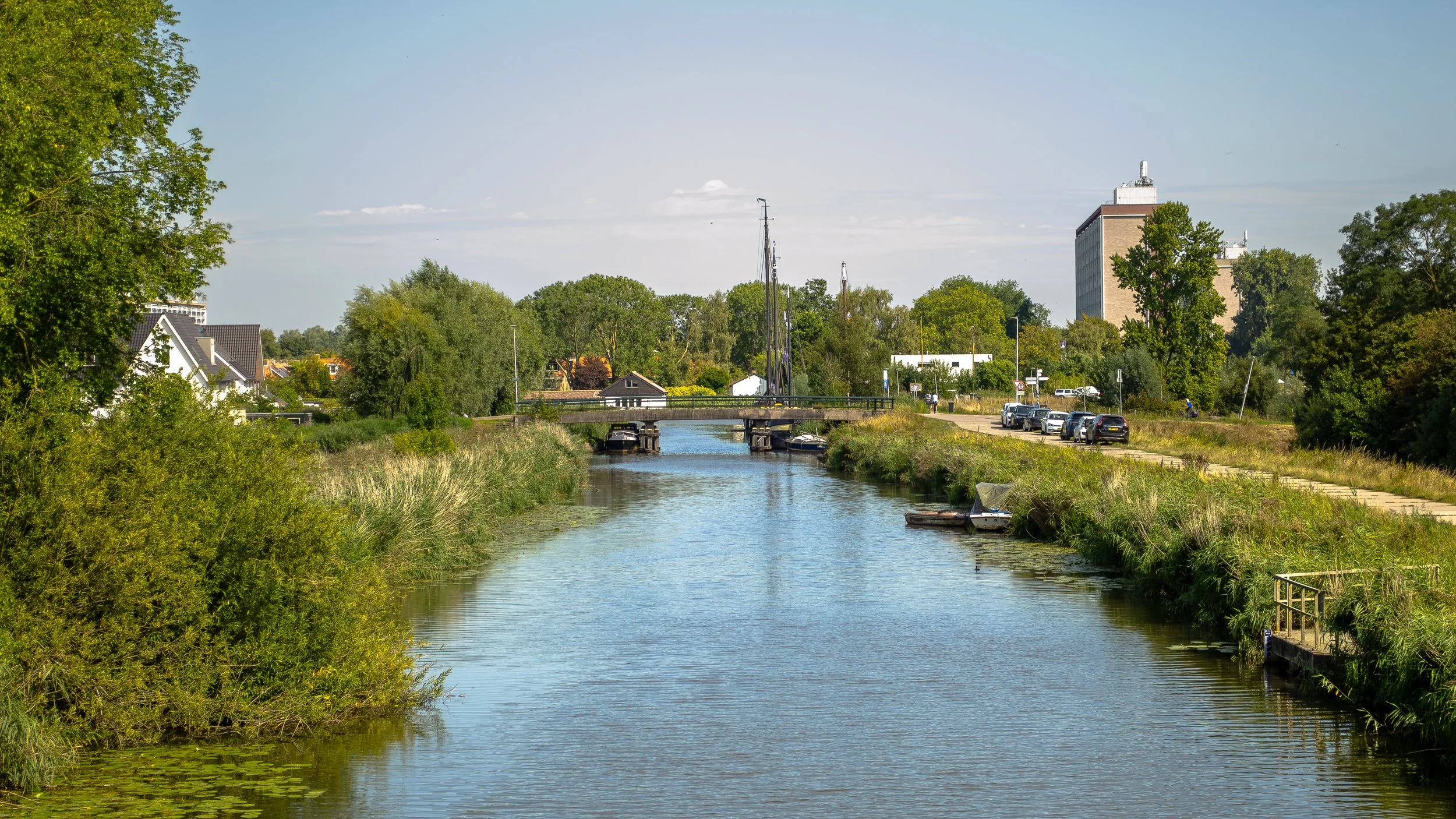 A calm river with a small boat docked by the grassy riverbank, a bridge crossing over, surrounded by green trees and a clear blue sky with some clouds, with buildings in the background.