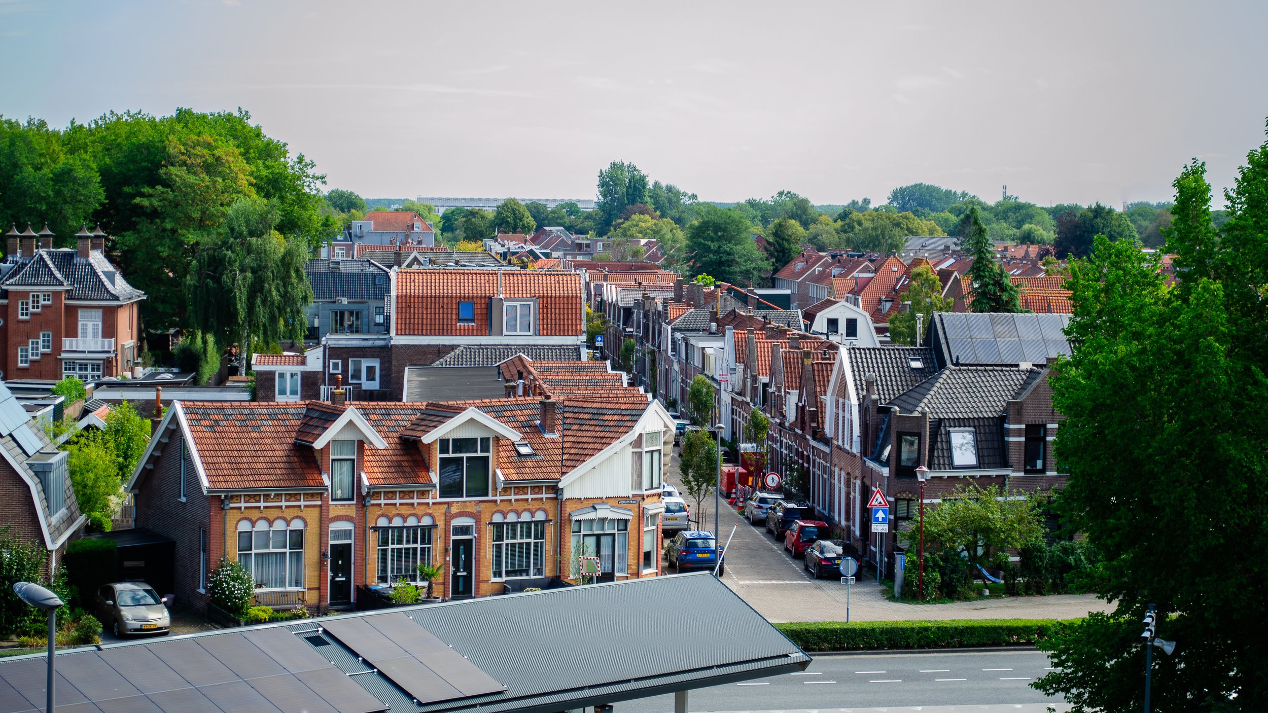 A view of a residential neighborhood with houses featuring red and gray tiled roofs, surrounded by lush green trees under a partly cloudy sky.