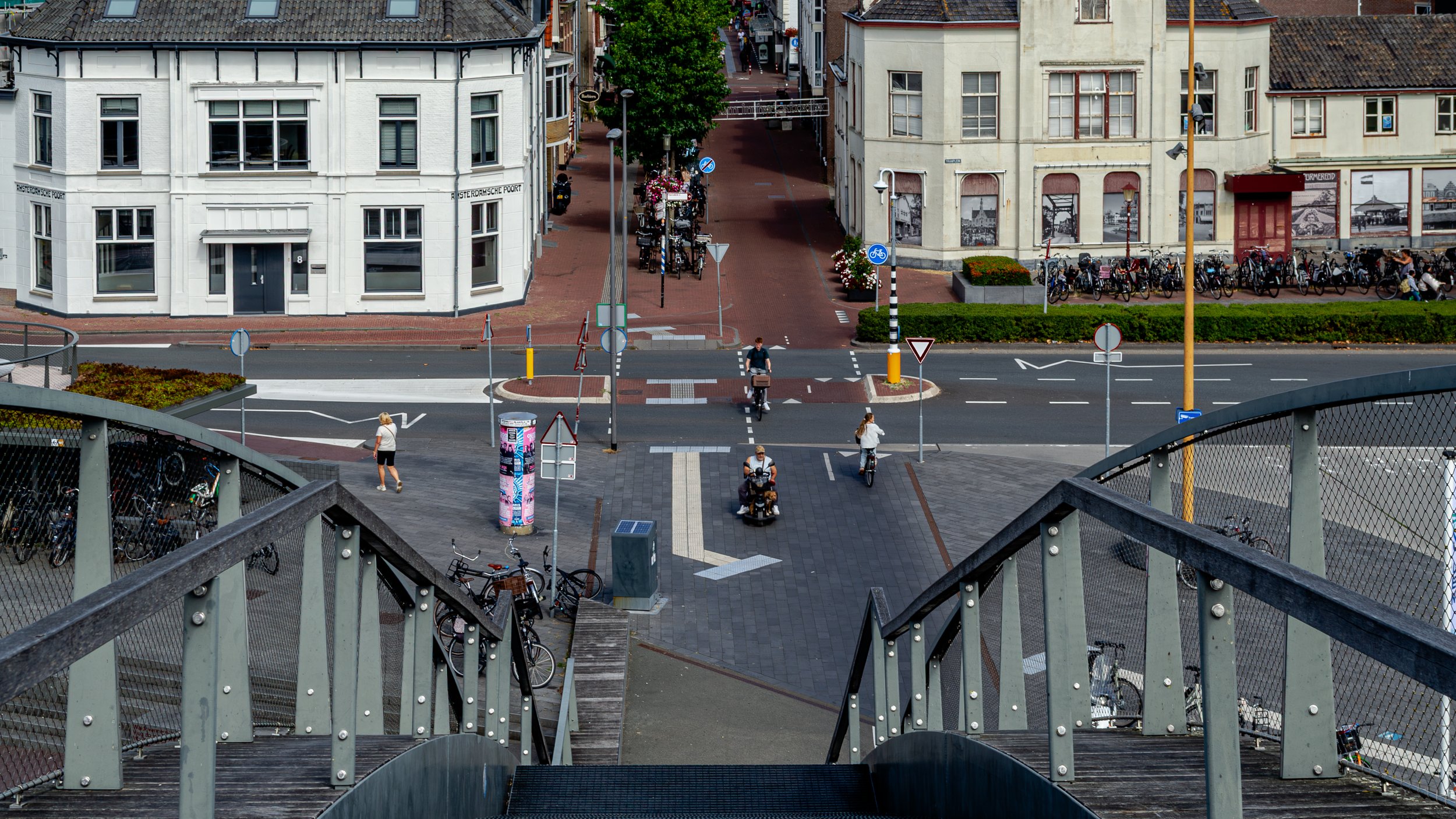 View from a staircase overlooking a city street with people, bicycles, and buildings, including a corner building with white and beige facades. The street has crossing lines, signs, and a few vehicles, with pedestrians walking and biking across.
