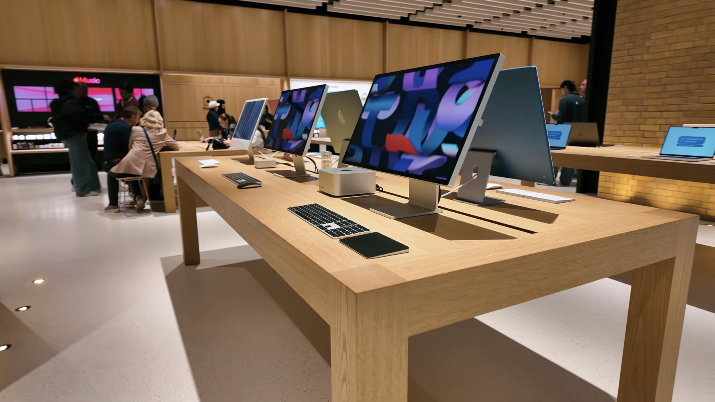 Several people browsing Apple iMac computers on display in an electronics store with wooden walls and desks.