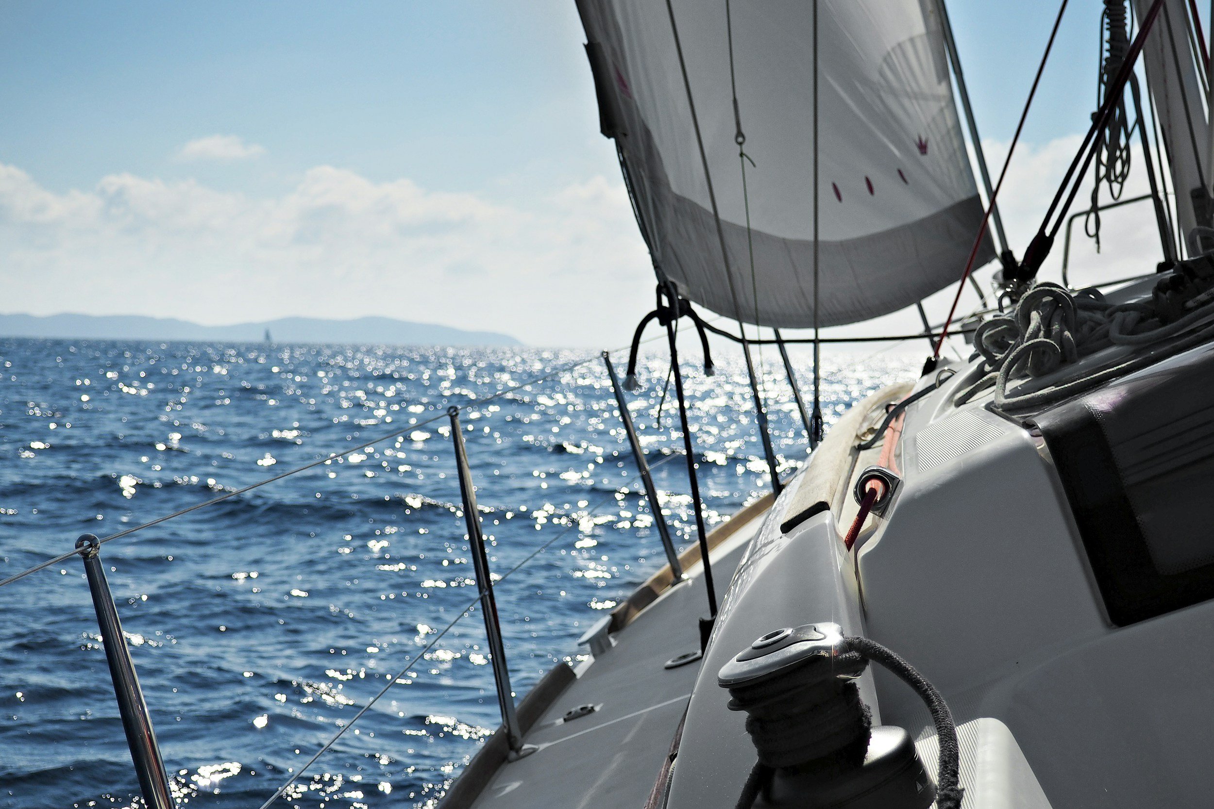 A view from a sailboat looking out over the water with a sail in the foreground and distant land on the horizon under a partly cloudy sky.