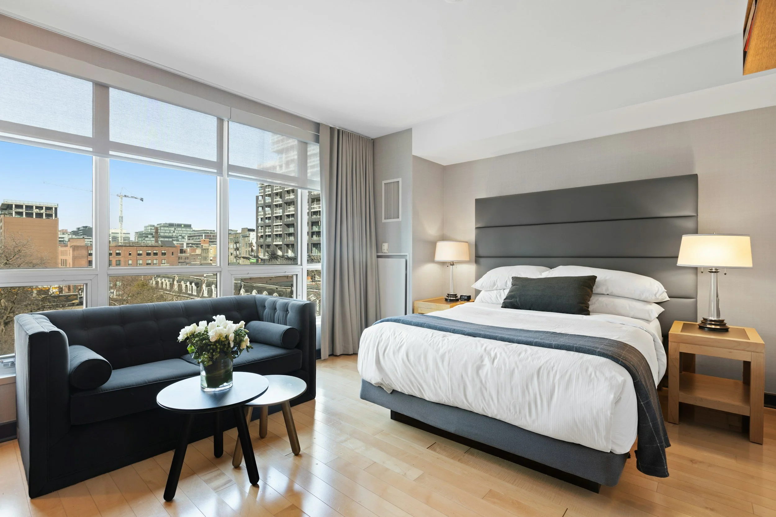 Hotel room with large window overlooking buildings; features a bed with white linens, black pillows, and dark headboard, nightstands with lamps, a small table with flowers, and a black sofa.